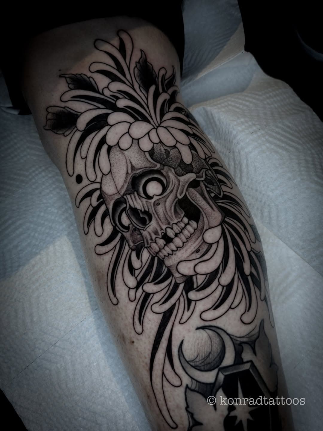 Black and grey illustrative tattoo of a skull adorned with ornate chrysanthemum flowers and decorative swirls on forearm