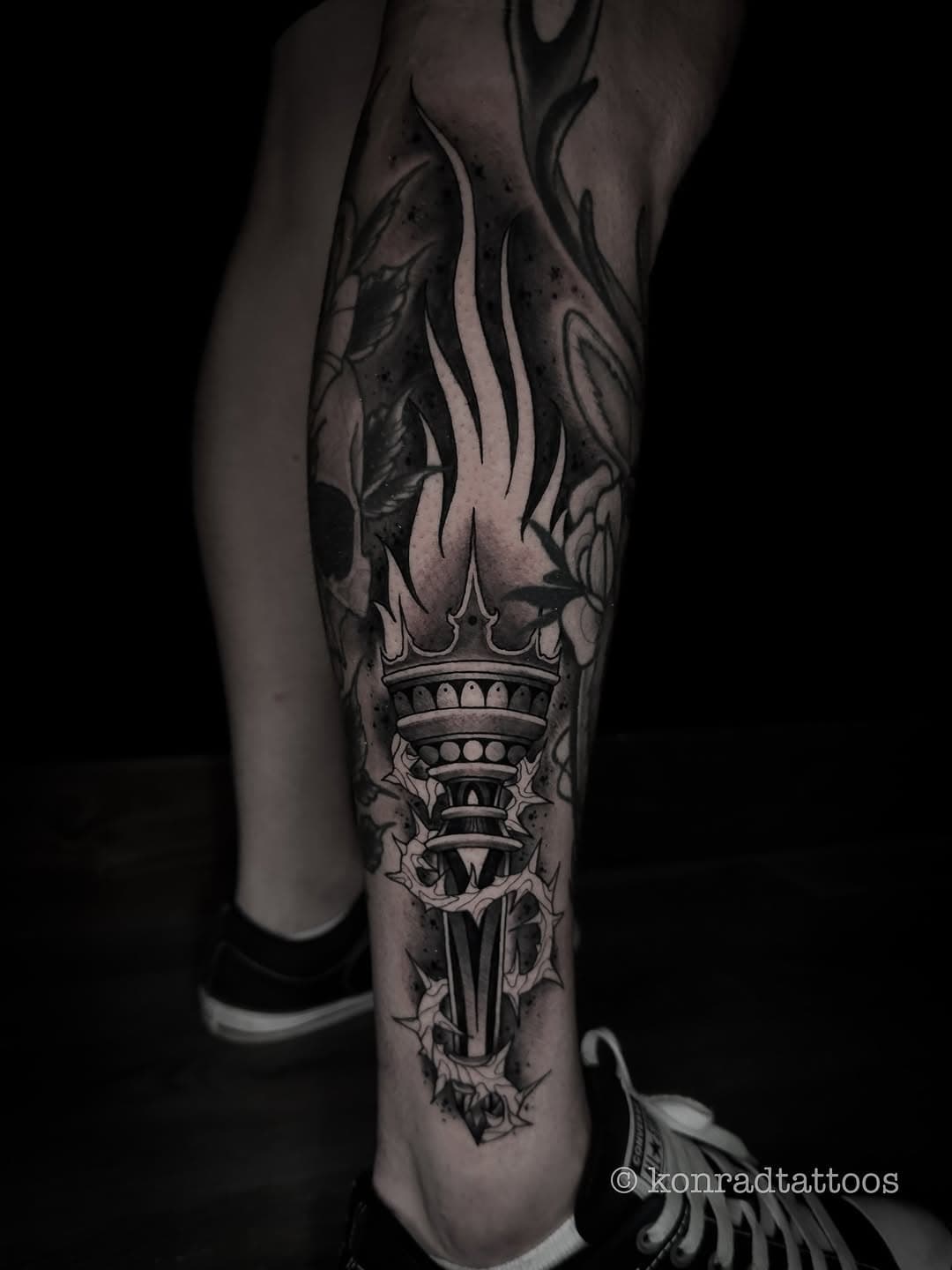 Black and grey leg tattoo featuring a flaming torch with ornate crown-like top surrounded by decorative leaves and flowing flames