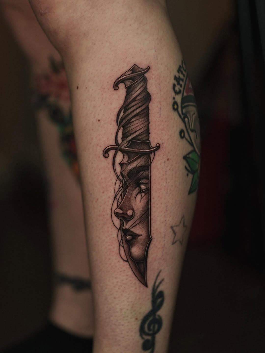 Black and grey illustrative tattoo of a dagger with a woman's face incorporated into the blade, done in dotwork style