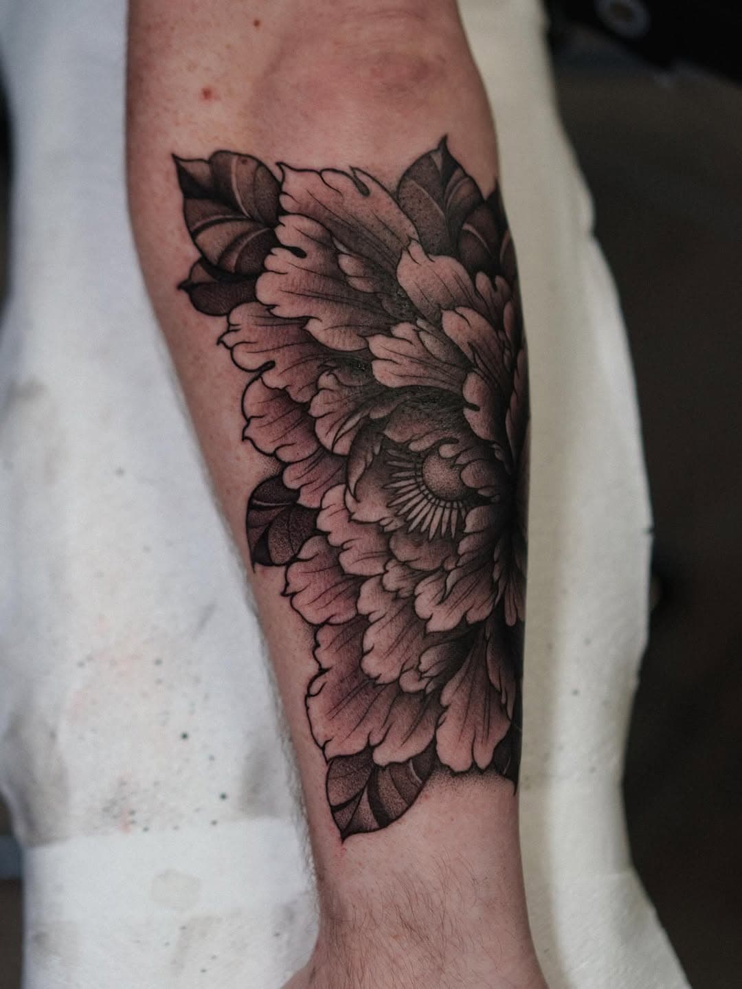 Black and grey illustrative peony flower tattoo with dotwork shading and leaves on forearm