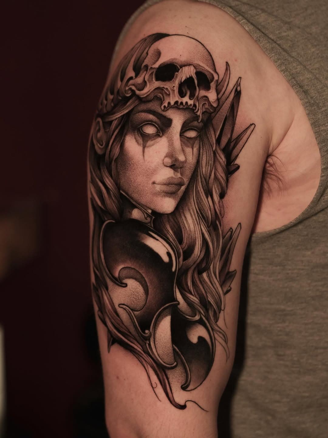 Black and grey illustrative tattoo of a woman's face with blank eyes wearing a skull headdress and ornate flowing details on upper arm