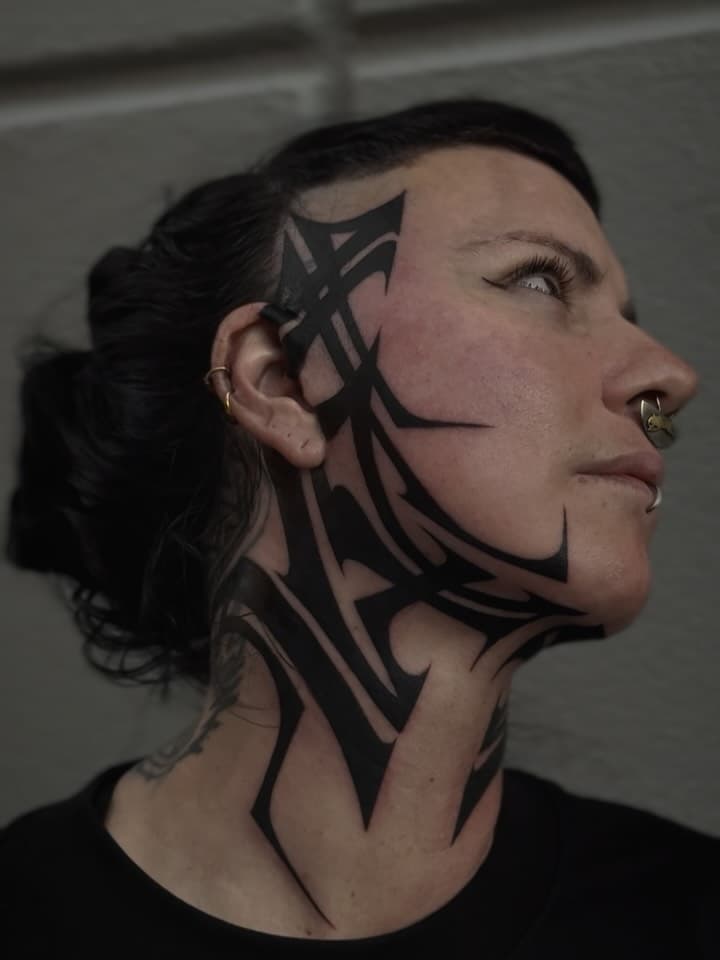 Bold blackwork tribal tattoo covering side of face and neck with geometric angular patterns and sharp pointed designs