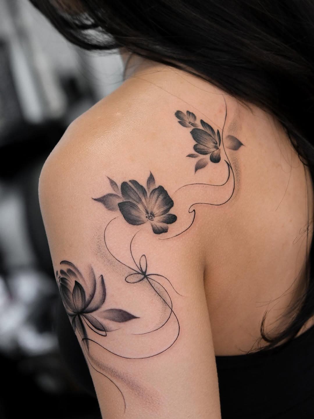Black and grey floral tattoo with three delicate flowers connected by flowing vine lines on shoulder and upper arm