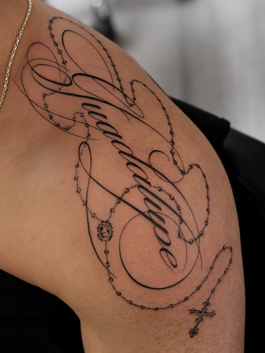 Shoulder tattoo featuring elegant script lettering intertwined with rosary beads and a cross in fine-line black ink