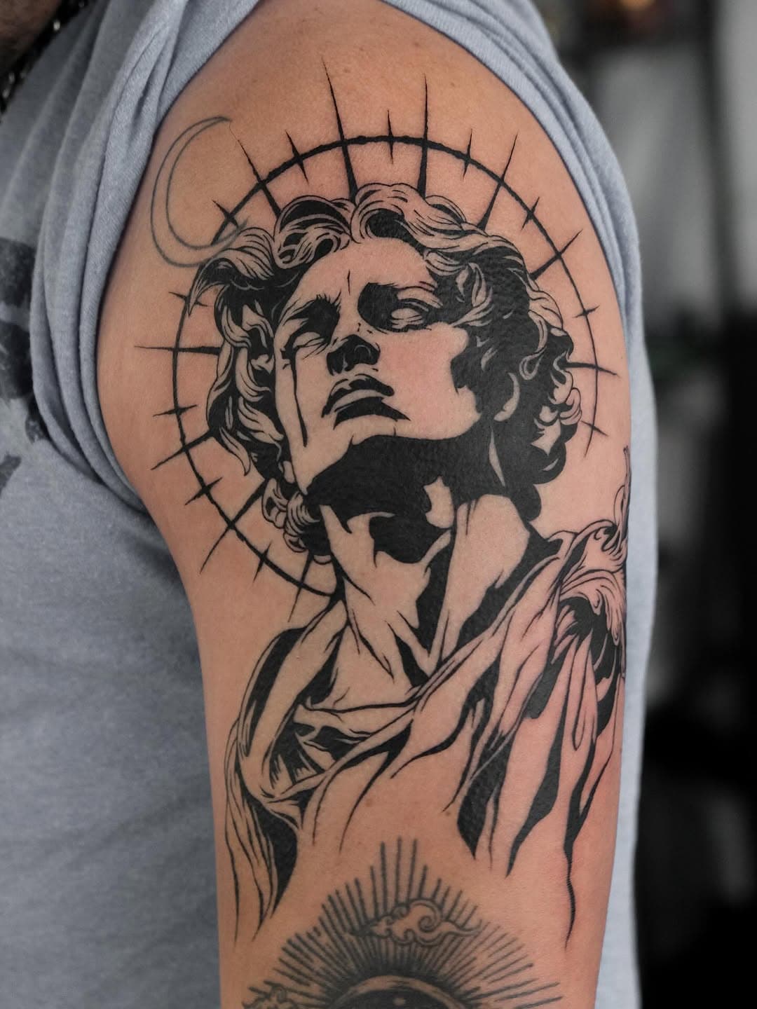 Black and grey tattoo of an angel with curly hair looking upward, featuring radiating halo lines and decorative drapery on upper arm