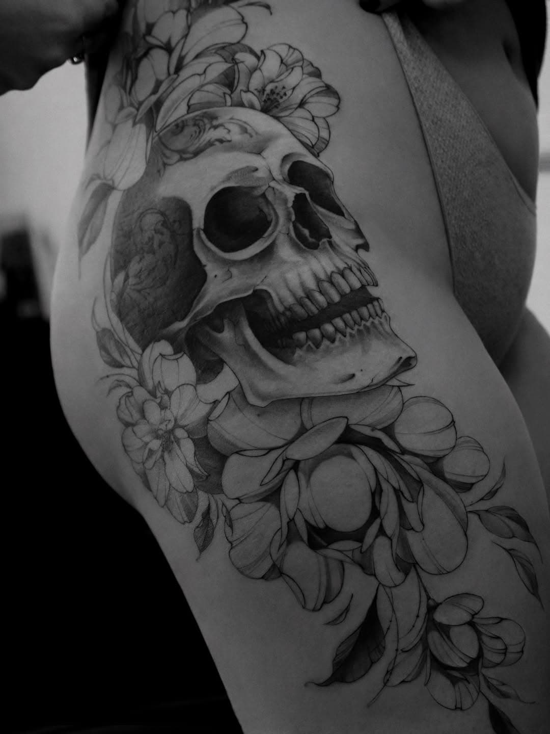 Black and grey thigh tattoo featuring a realistic human skull surrounded by delicate floral blooms and botanical elements