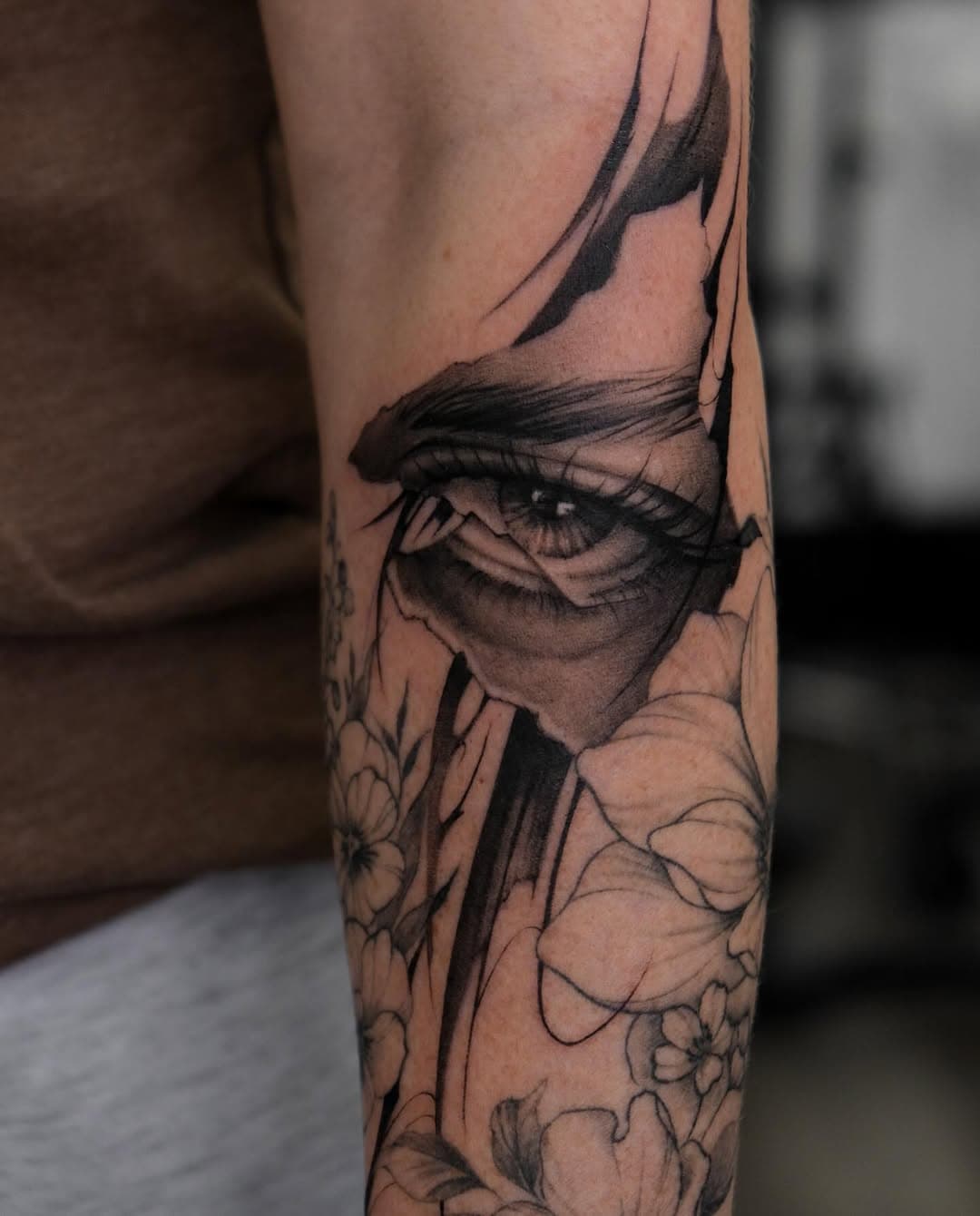 Black and grey realistic forearm tattoo featuring a detailed eye with dramatic shading surrounded by delicate floral elements