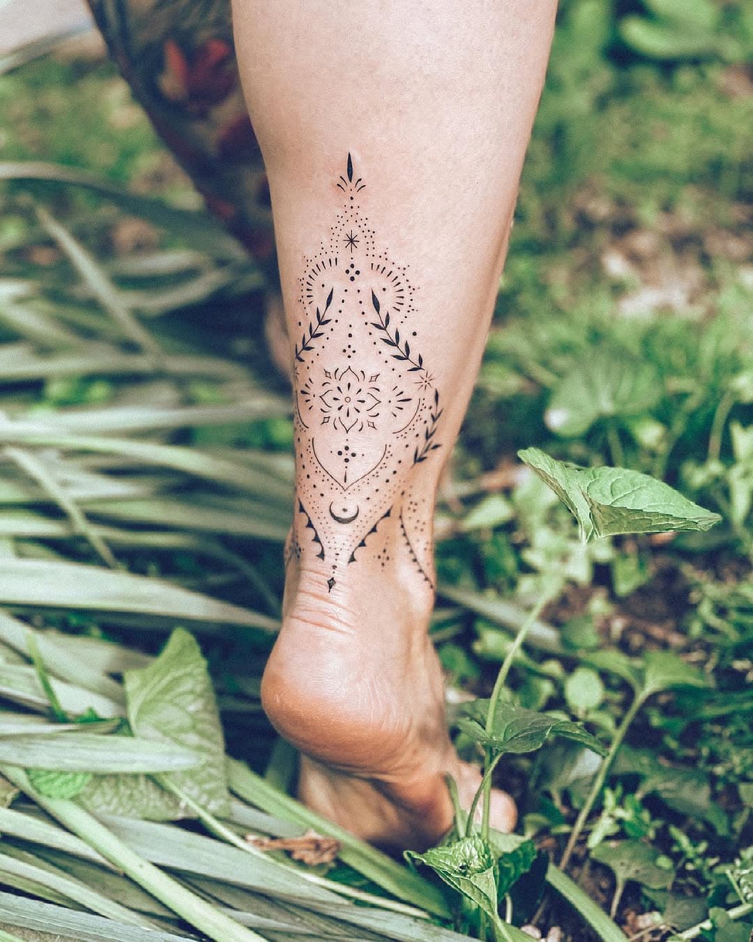 Delicate dotwork tattoo on back of leg featuring mandala flower, crescents, leaves, and geometric patterns in black ink