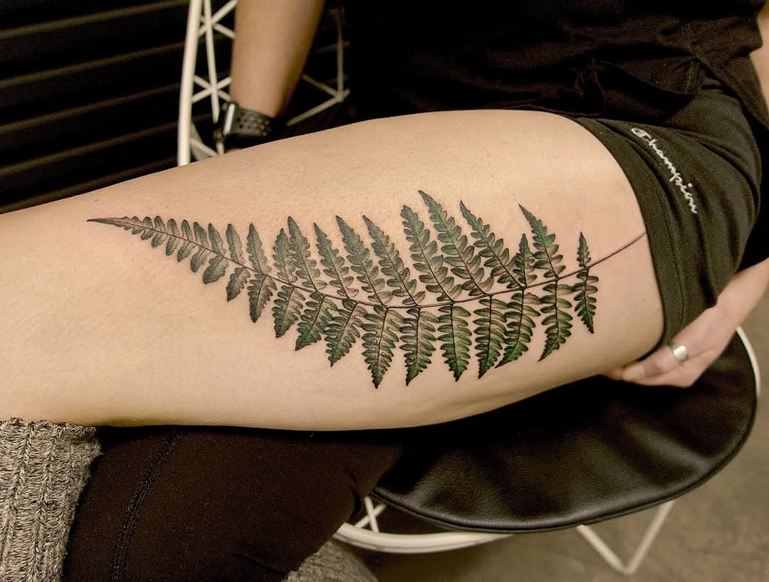 Detailed fern leaf tattoo in green and brown tones on forearm, featuring realistic botanical illustration with intricate fronds