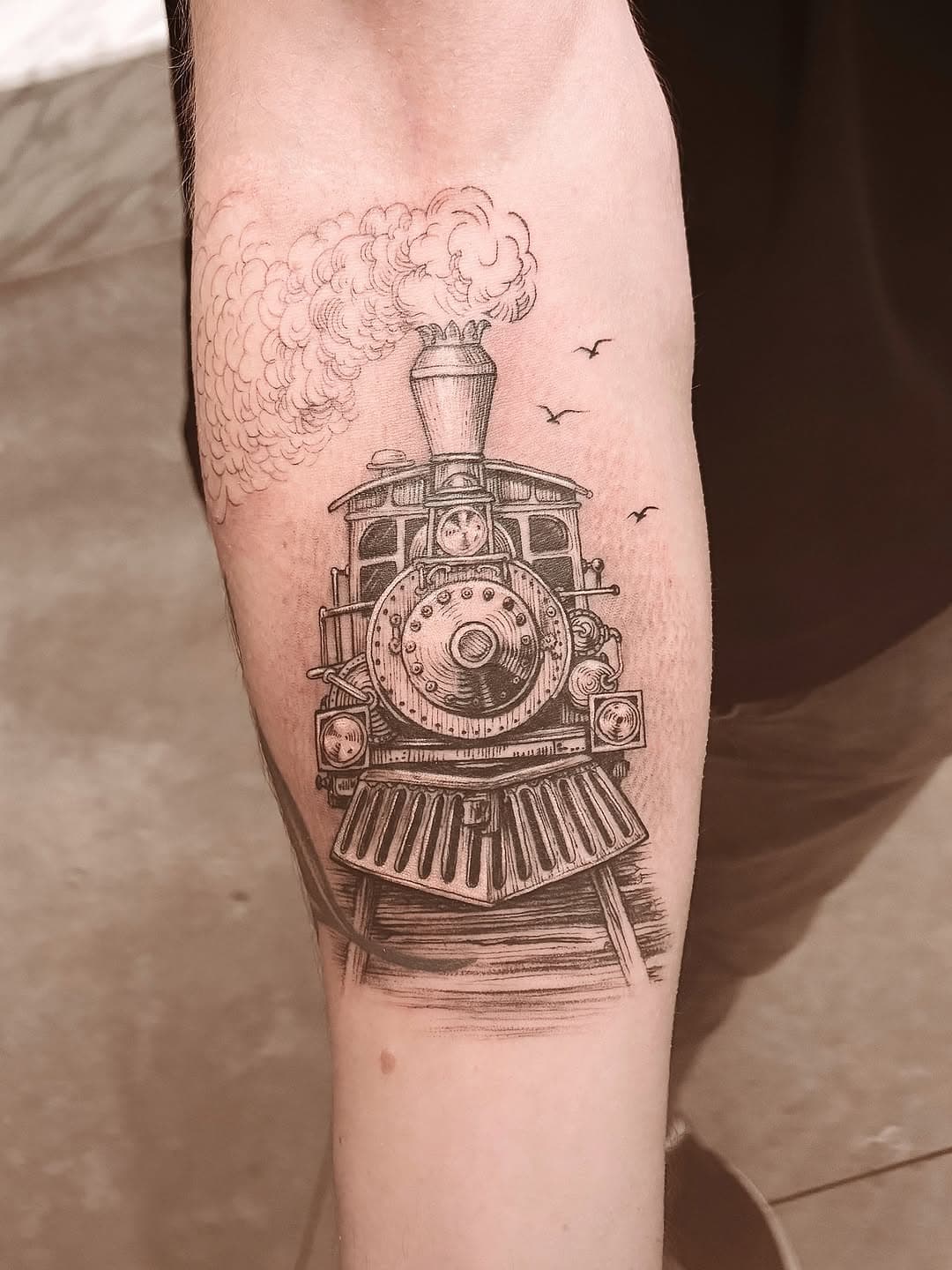 Detailed vintage steam locomotive tattoo on forearm with billowing smoke and flying birds in fine line illustrative style