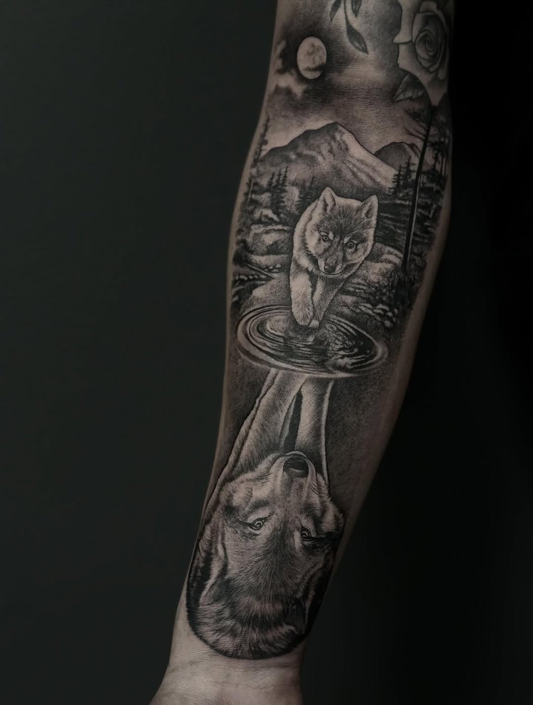 Black and grey realism sleeve tattoo featuring wolves in nature scene with mountains, water ripples, and forest elements