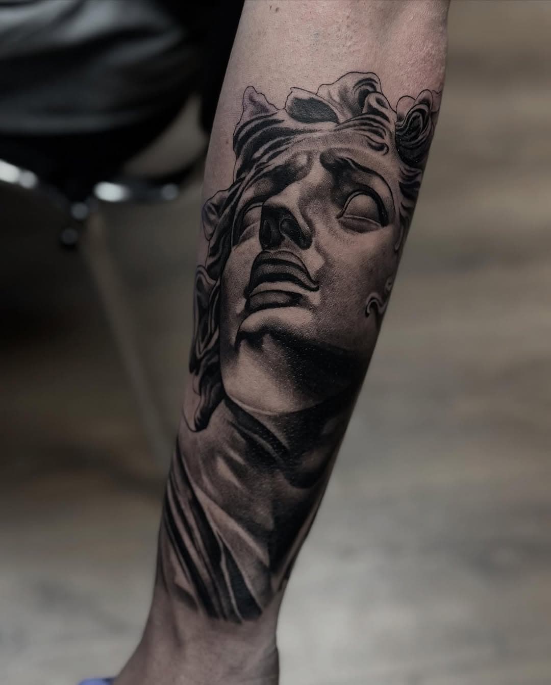 Black and grey realistic tattoo of a classical Greek or Roman statue bust with crown, featuring detailed shading on forearm