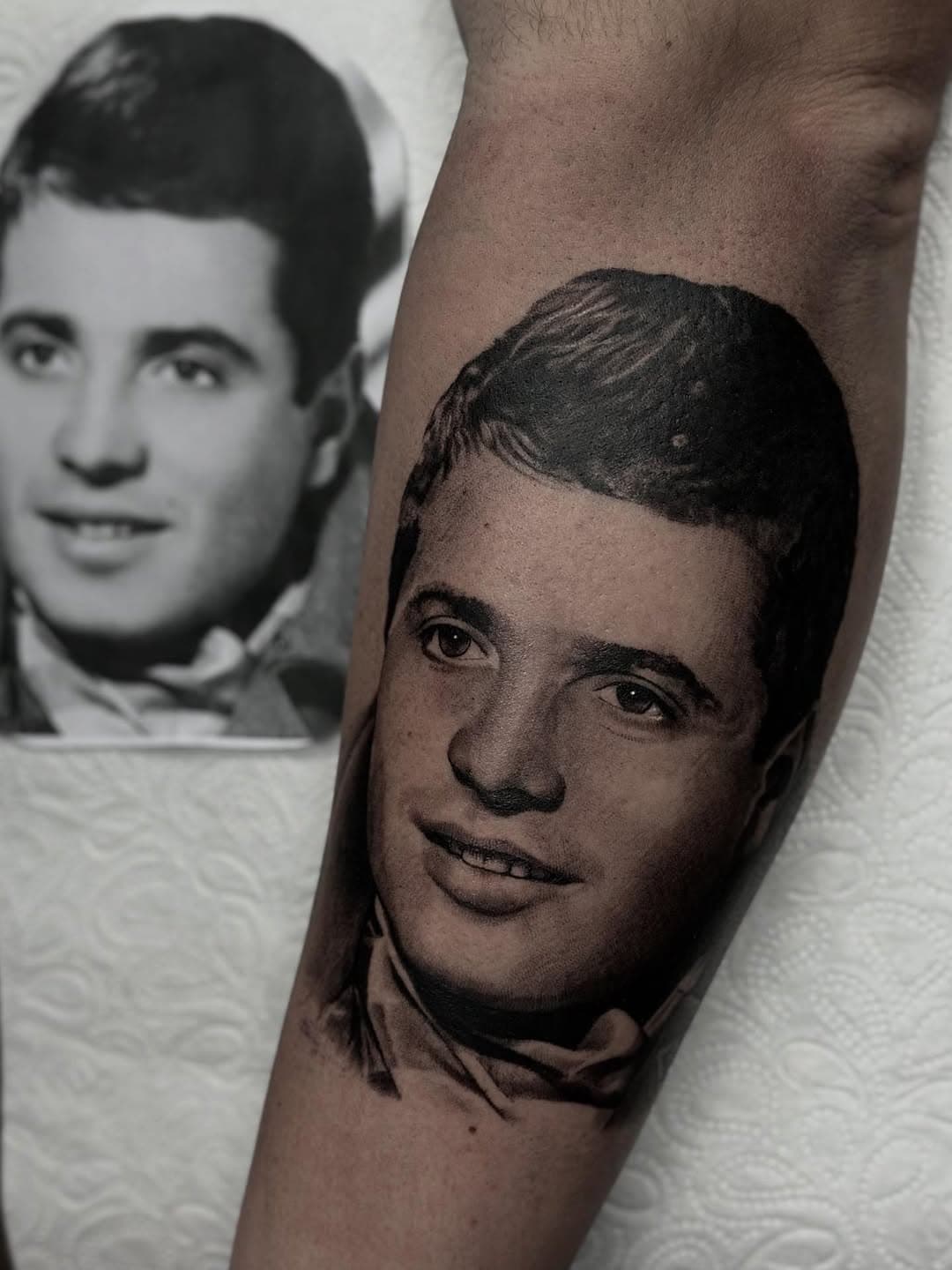 Realistic black and grey portrait tattoo of a young man's face on forearm, based on vintage photograph shown beside it