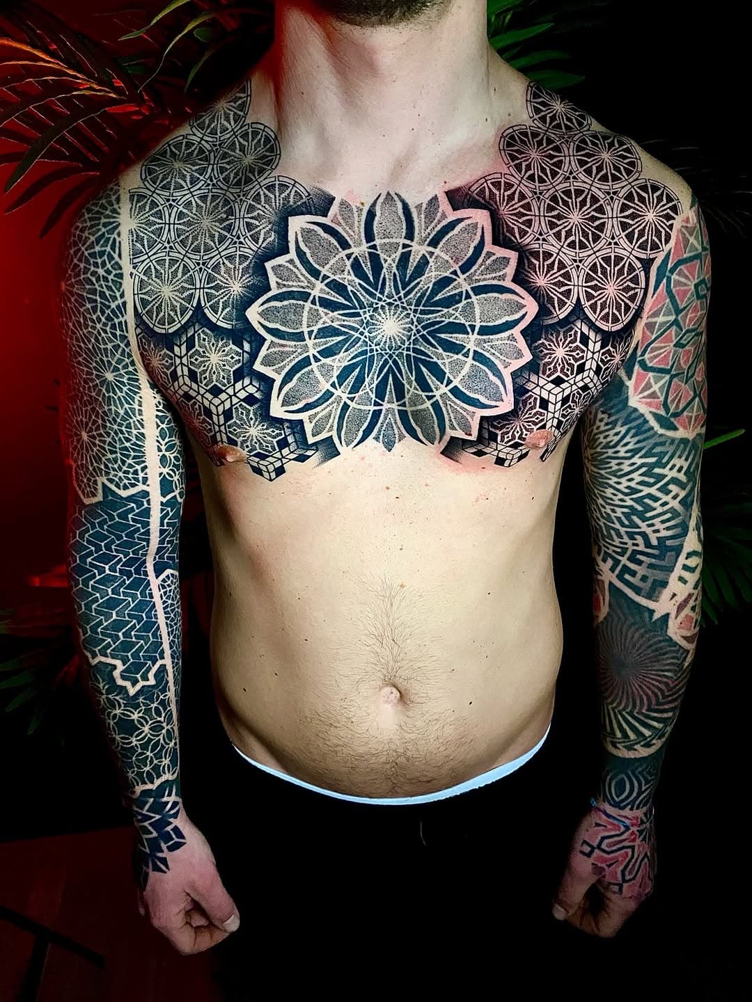 Intricate geometric blackwork tattoo featuring mandala flower centerpiece with sacred geometry patterns across chest, shoulders and full sleeves
