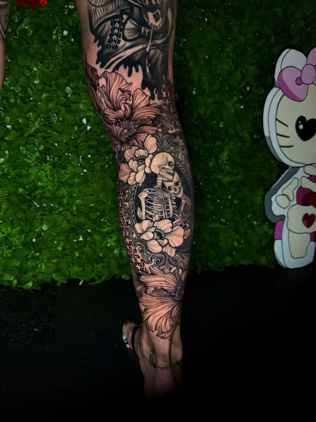 Black-and-grey leg sleeve tattoo featuring ornate flowers, skeleton, and decorative baroque elements in illustrative blackwork style
