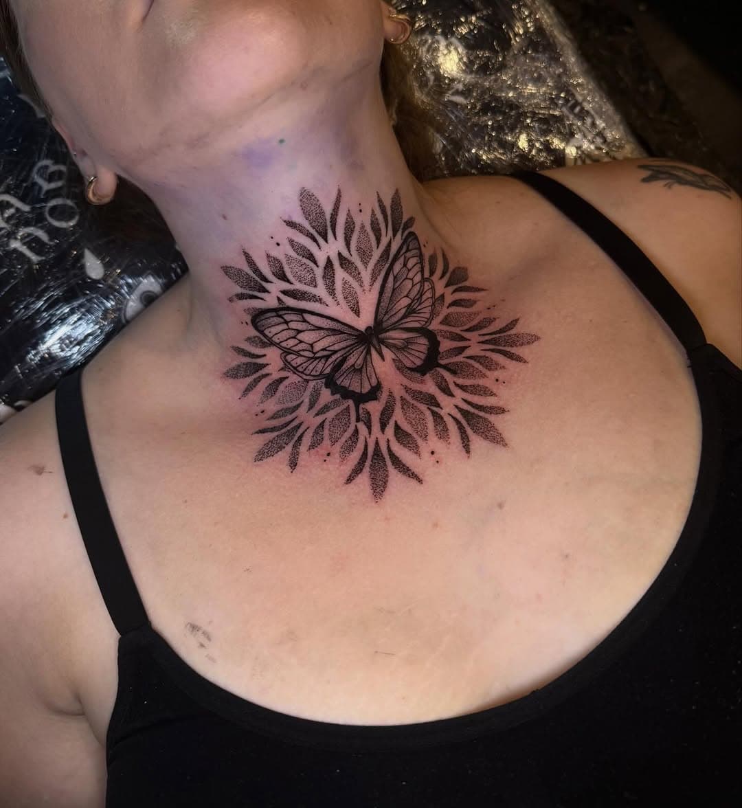 Black-and-grey butterfly tattoo with radiating botanical leaves and dotwork details on upper chest and collarbone area