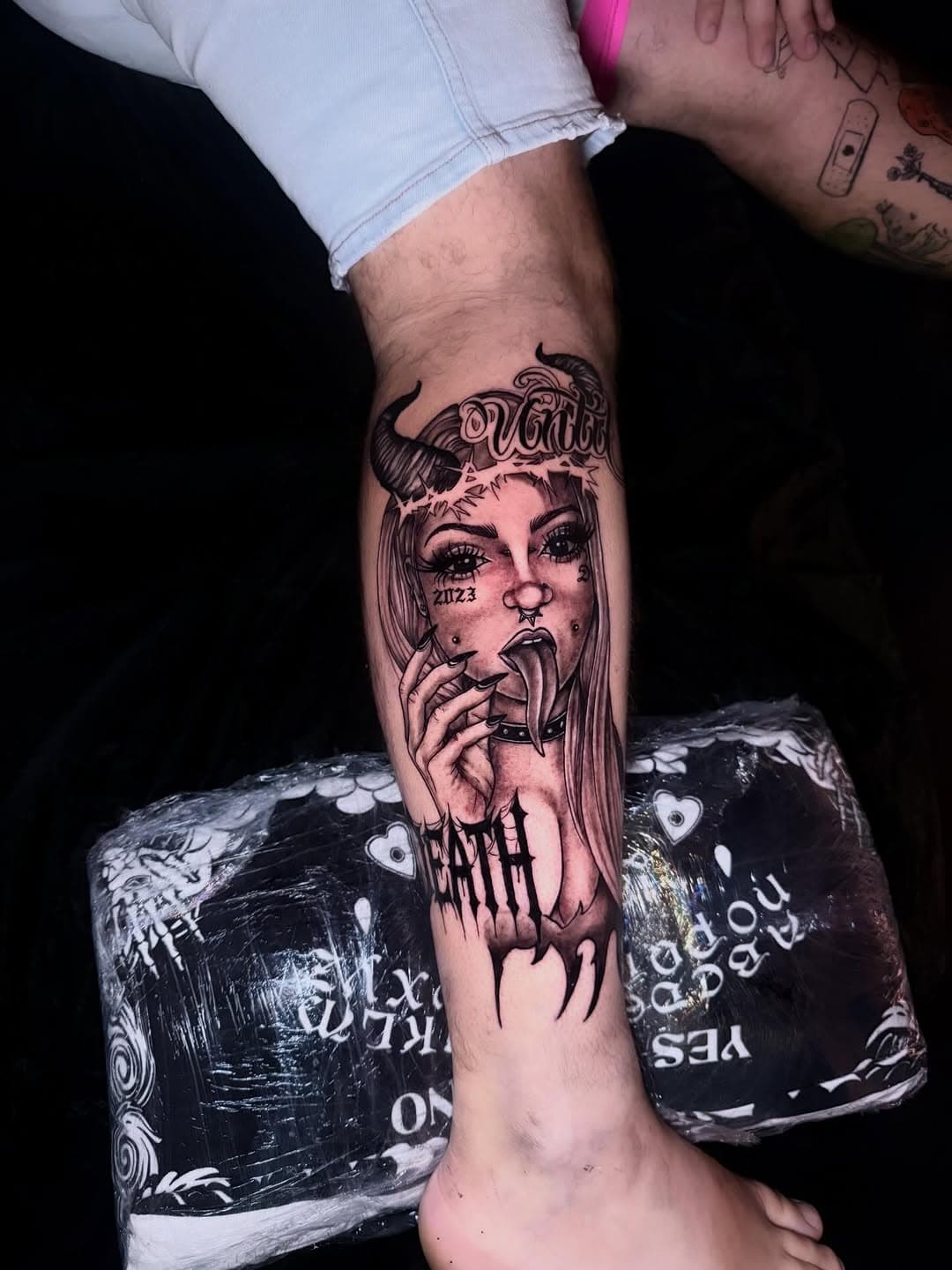 Black-and-grey illustrative tattoo of a stylized woman's face with crown, dramatic makeup, wearing headphones, with dripping ink effects on forearm