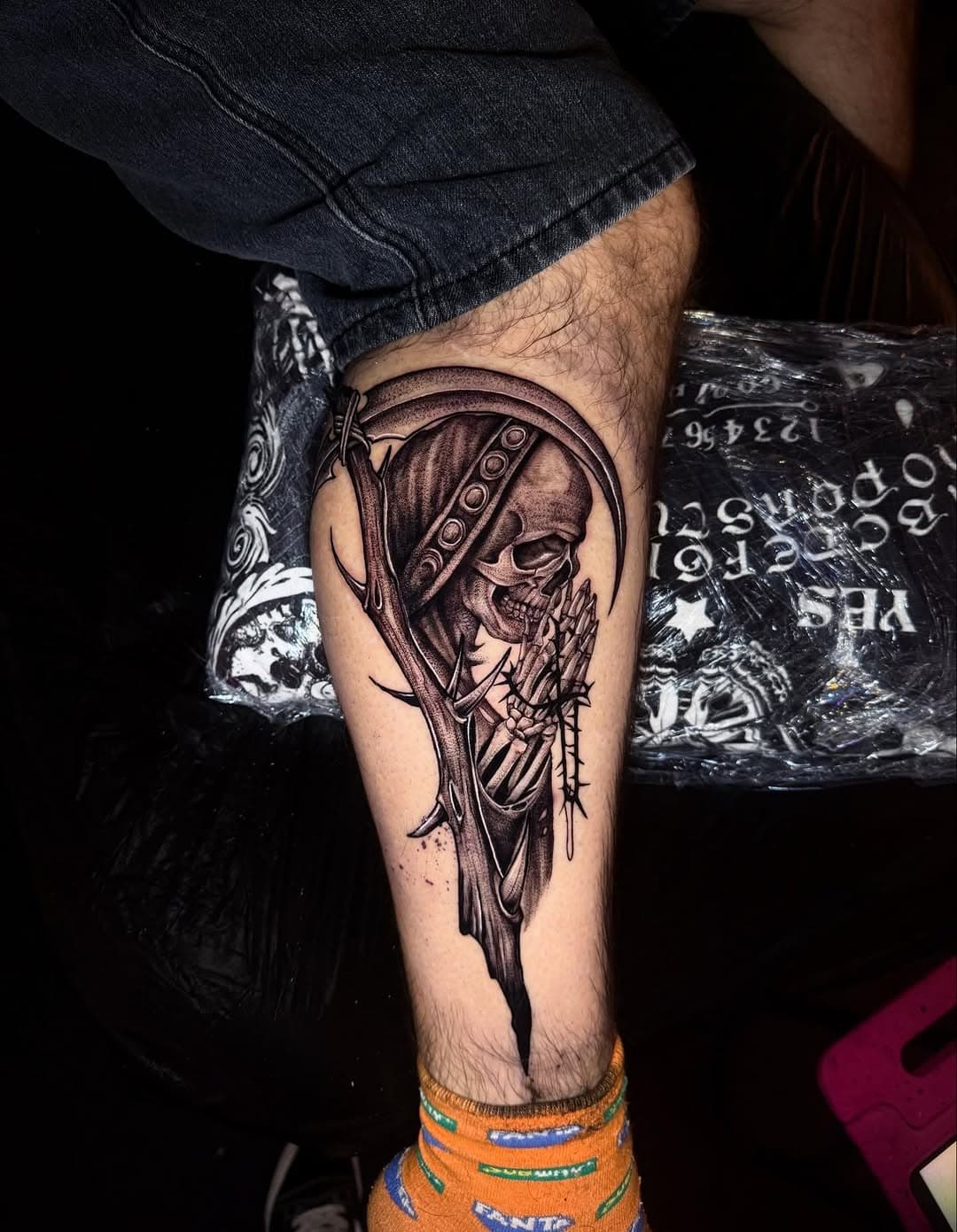 Black and grey grim reaper tattoo with skull face holding scythe on lower leg, featuring illustrative blackwork style with dripping ink effects