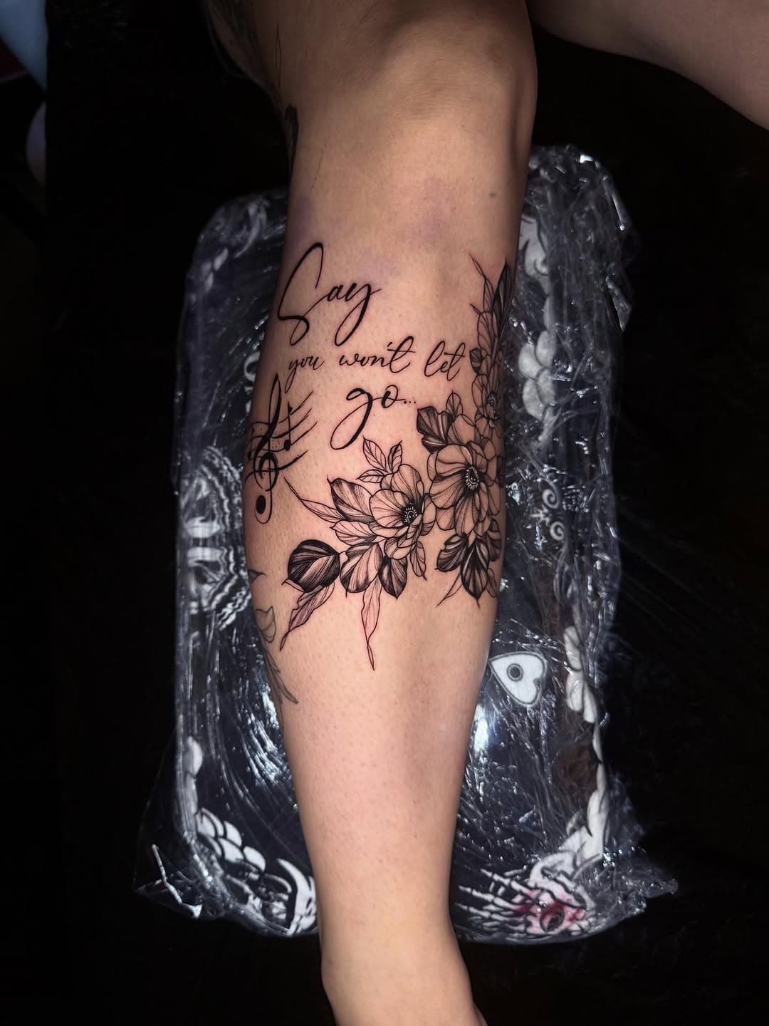 Black and grey floral tattoo on forearm with script text reading Say you won't let go, featuring detailed botanical flowers and musical note elements