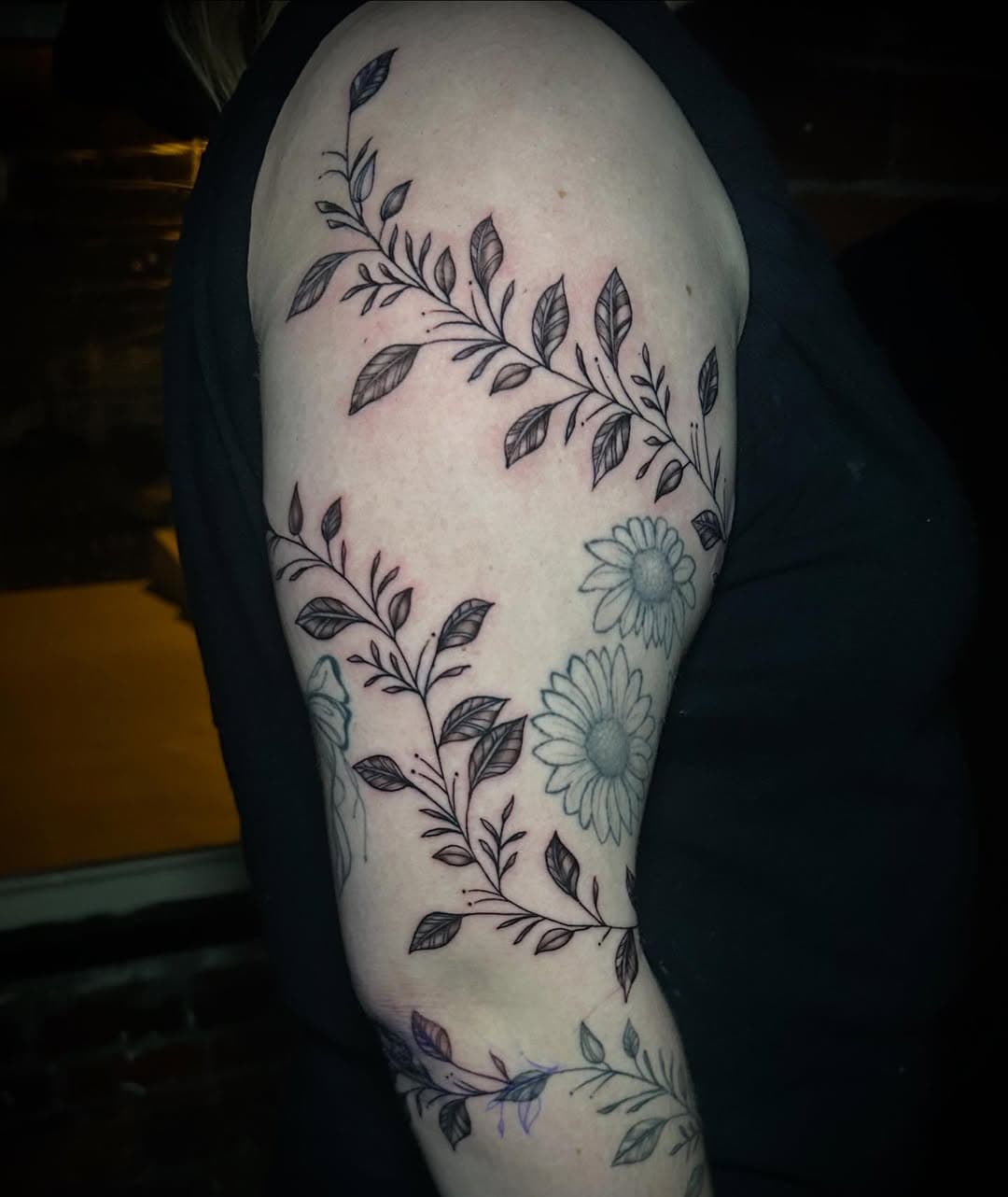 Black and grey illustrative tattoo of flowing botanical branches with leaves and blue-tinted daisies wrapping around upper arm and thigh
