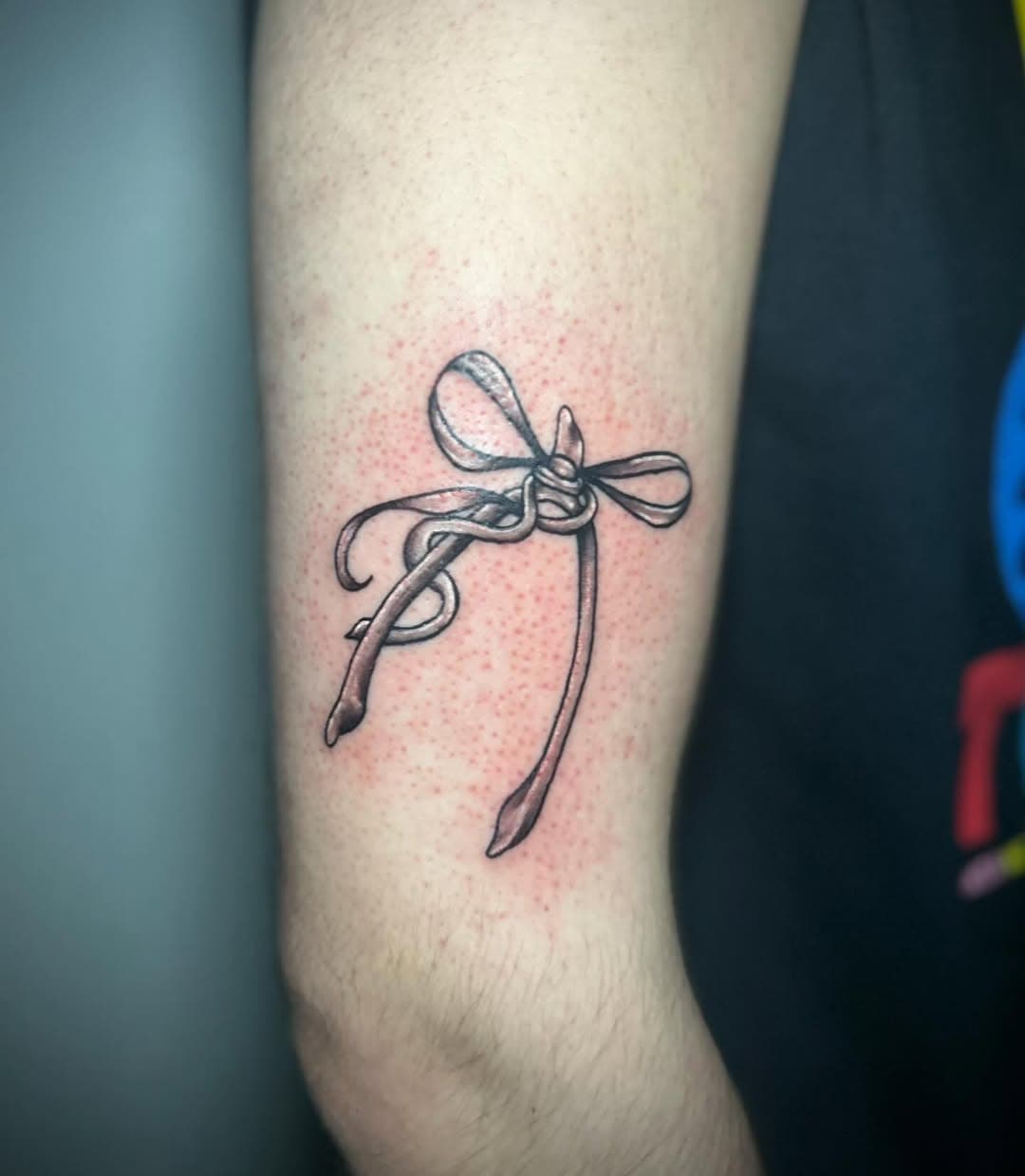 Black and grey illustrative tattoo of a delicate bow tied around the forearm, appearing like a ribbon wrapped around a gift
