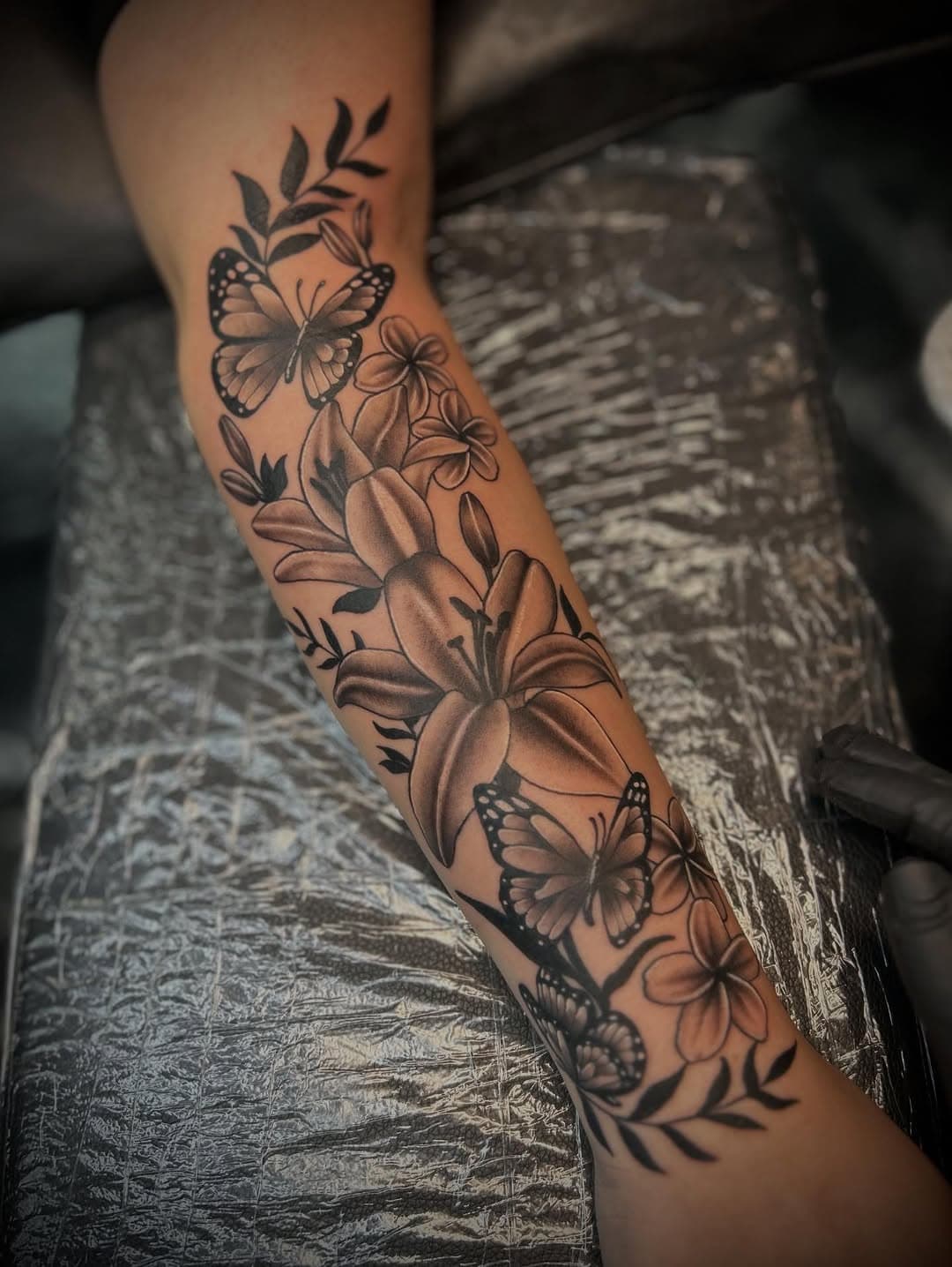 Black and grey forearm tattoo featuring butterflies, lily flowers, smaller blooms, and delicate leaf branches in illustrative style
