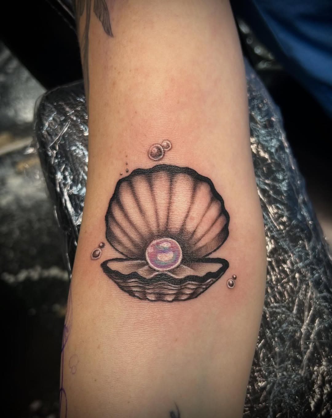 Black and grey illustrative tattoo of an open clamshell with an iridescent pearl inside and small bubbles floating above