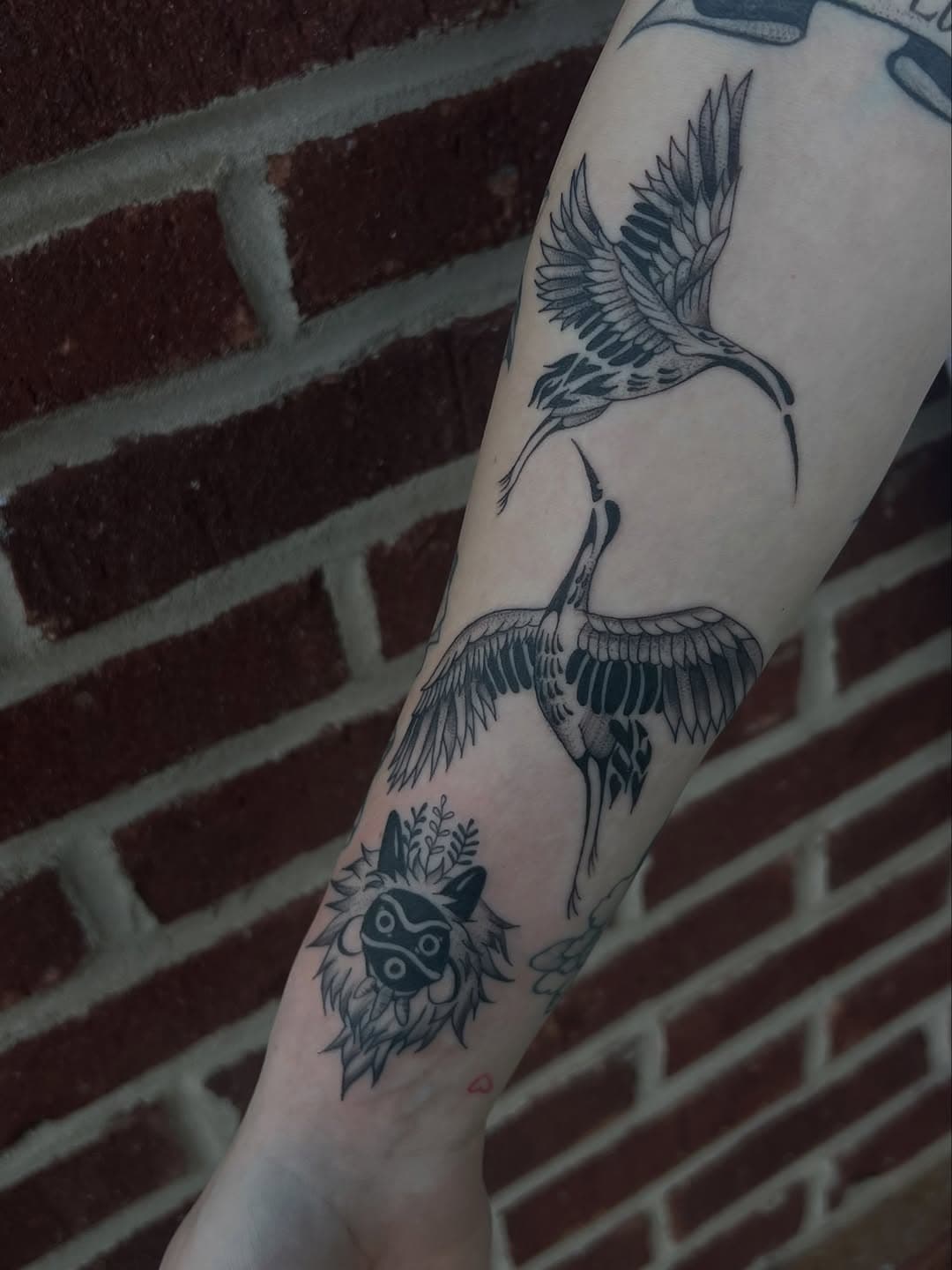 Black and grey leg tattoo featuring two cranes in flight and a stylized spirit mask with foliage in illustrative style