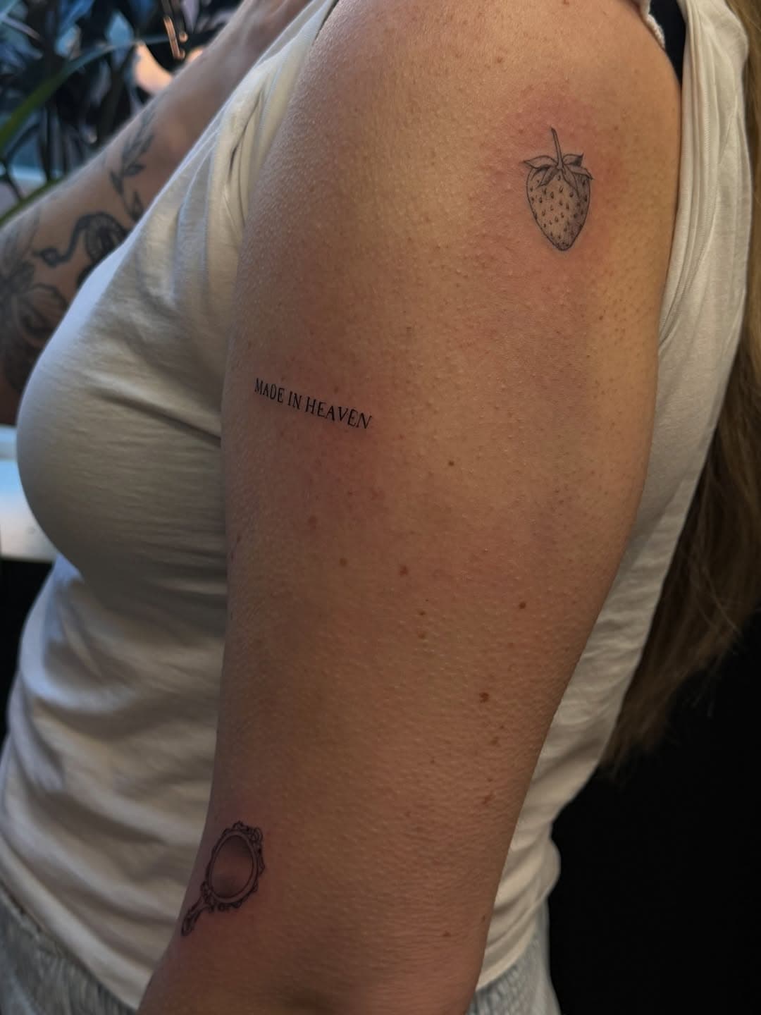 Black and grey tattoos on arm showing a strawberry at top, text reading MADE IN HEAVEN in middle, and an ornate mirror frame at bottom