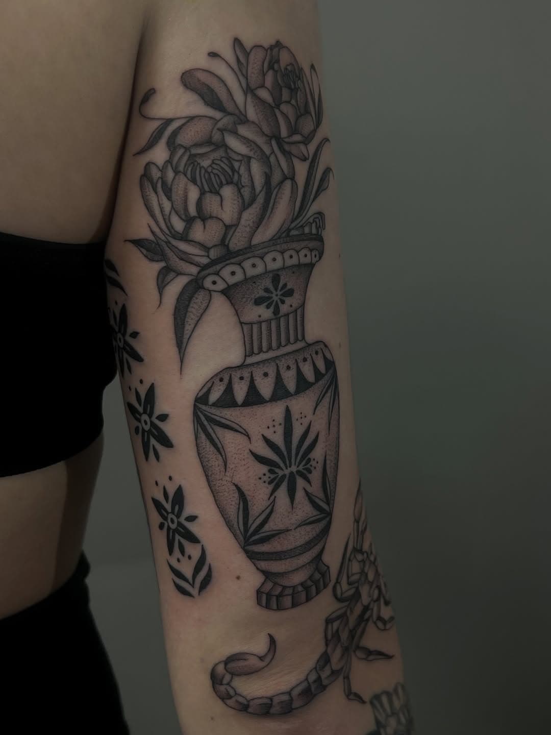 Black and grey illustrative tattoo of an ornate decorative vase with floral patterns containing blooming peonies and a snake below