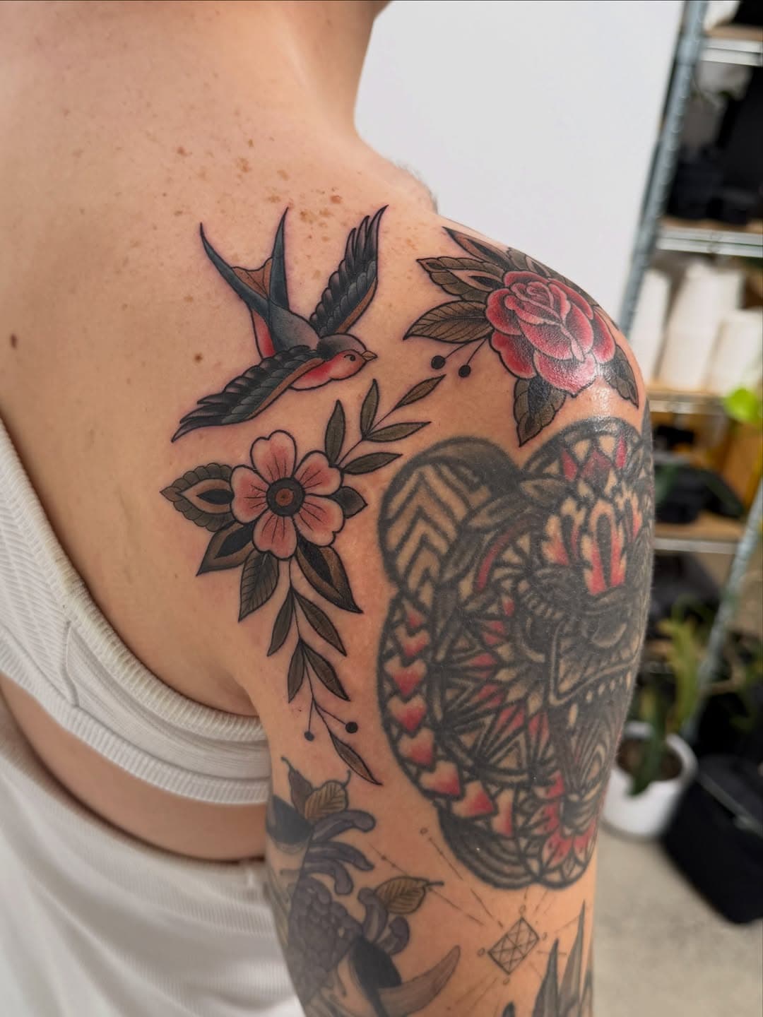 Traditional style tattoo featuring a black swallow bird, pink rose, and pink flower with leaves across shoulder and upper arm