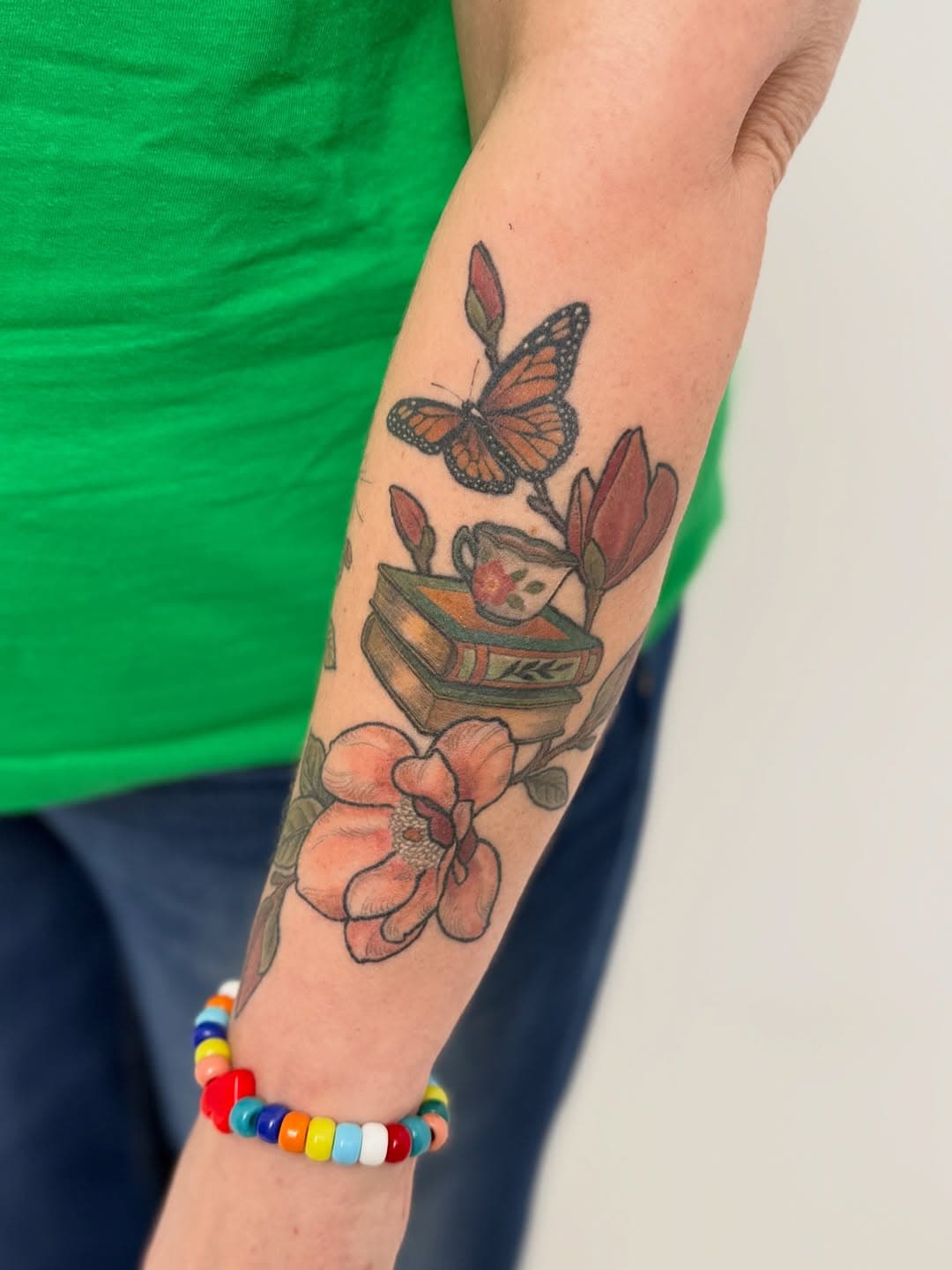 Colorful forearm tattoo featuring a monarch butterfly, stacked books with teacup, magnolia flowers and buds in illustrative neo-traditional style