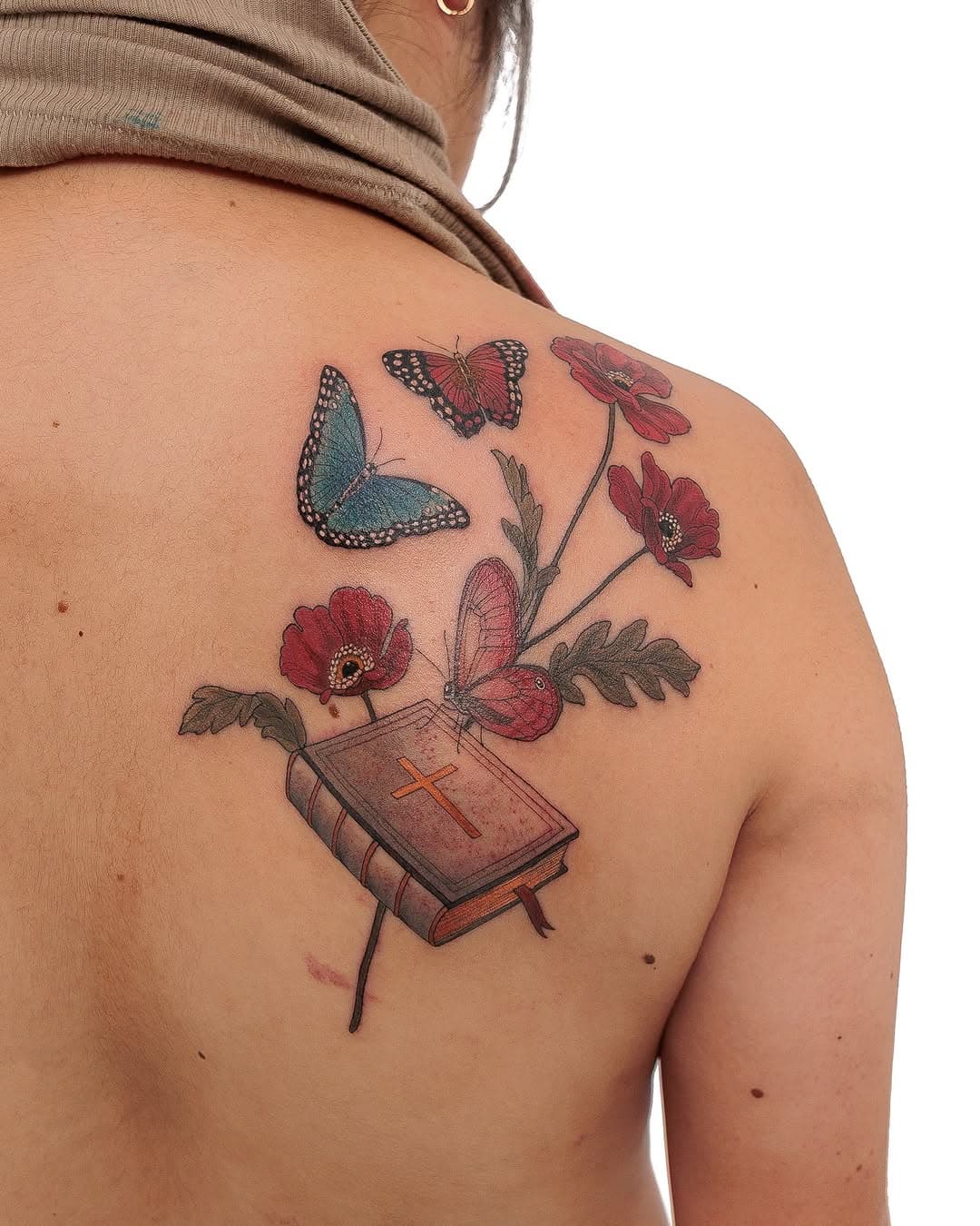 Illustrative back tattoo featuring a Bible with cross, red flowers, foliage, and three butterflies in teal and pink tones