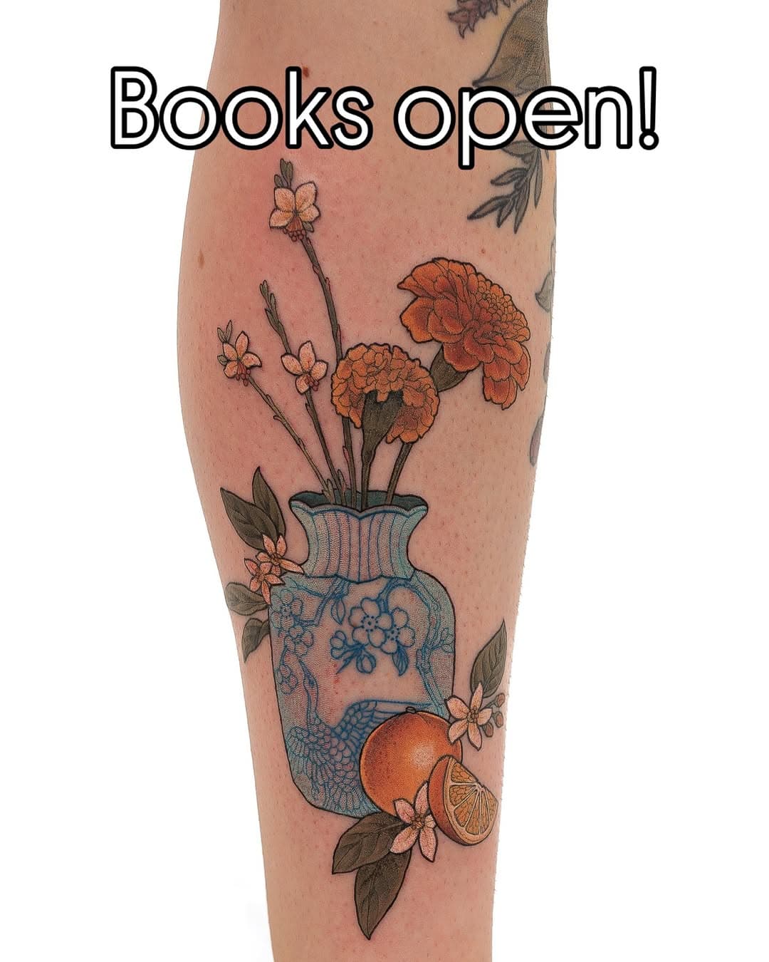 Illustrative leg tattoo featuring a blue decorative vase with orange marigolds, delicate branch flowers, citrus fruit slice, and green leaves