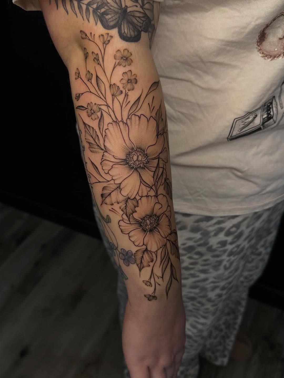 Black and grey floral sleeve tattoo featuring large poppies, delicate wildflowers, and botanical elements in fine-line illustrative style