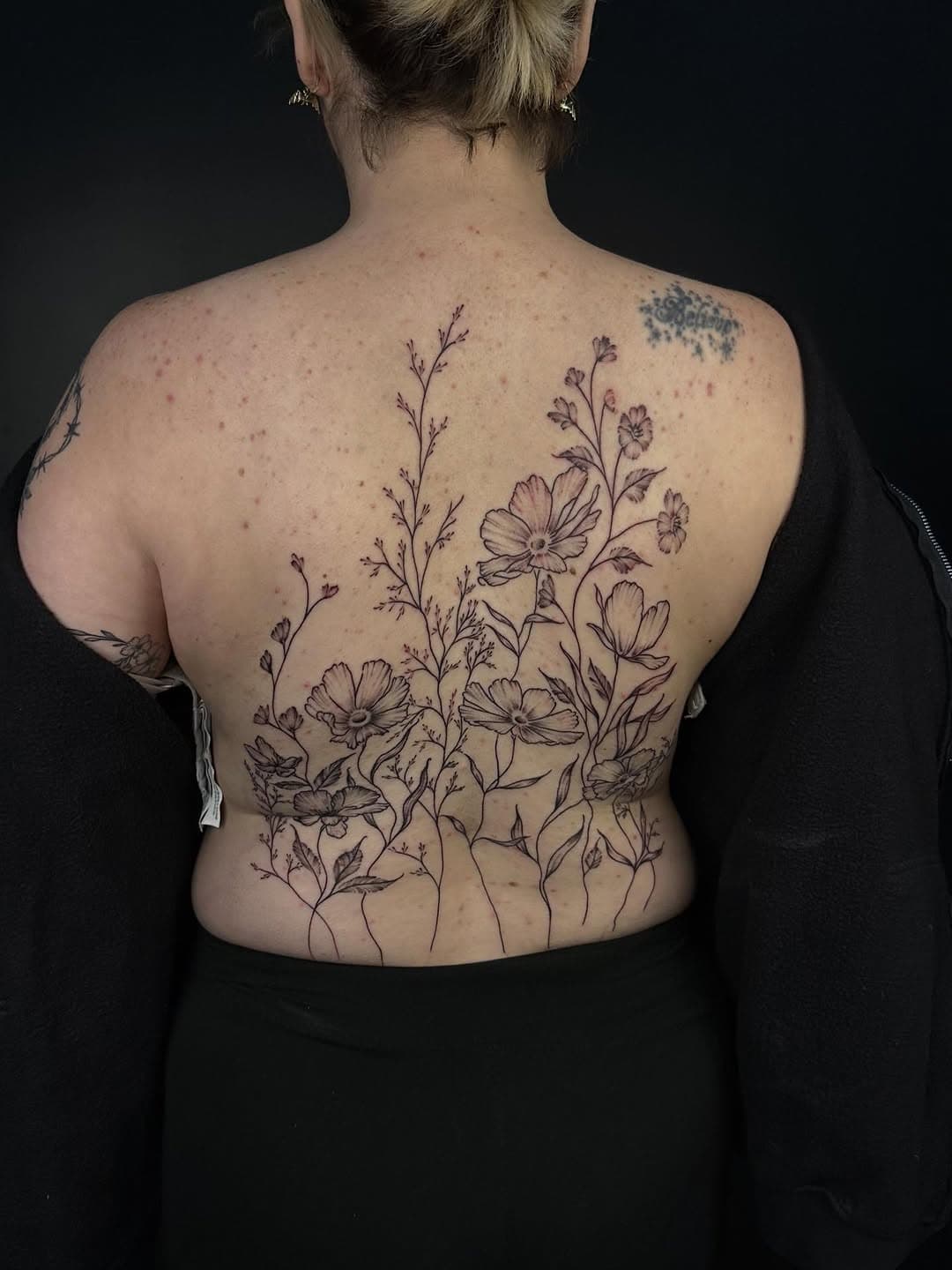 Black-and-grey fine-line botanical back tattoo featuring delicate wildflowers with cosmos blooms and stems in illustrative style