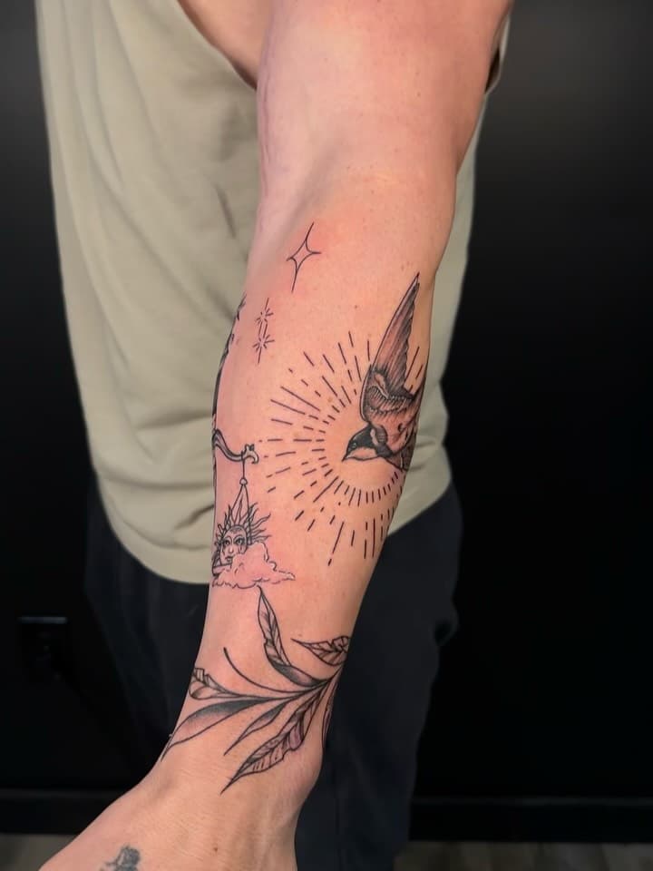 Black-and-grey illustrative forearm tattoo featuring a swallow bird in flight, radiating sun rays, celestial stars, crescent moon, and flowing feather