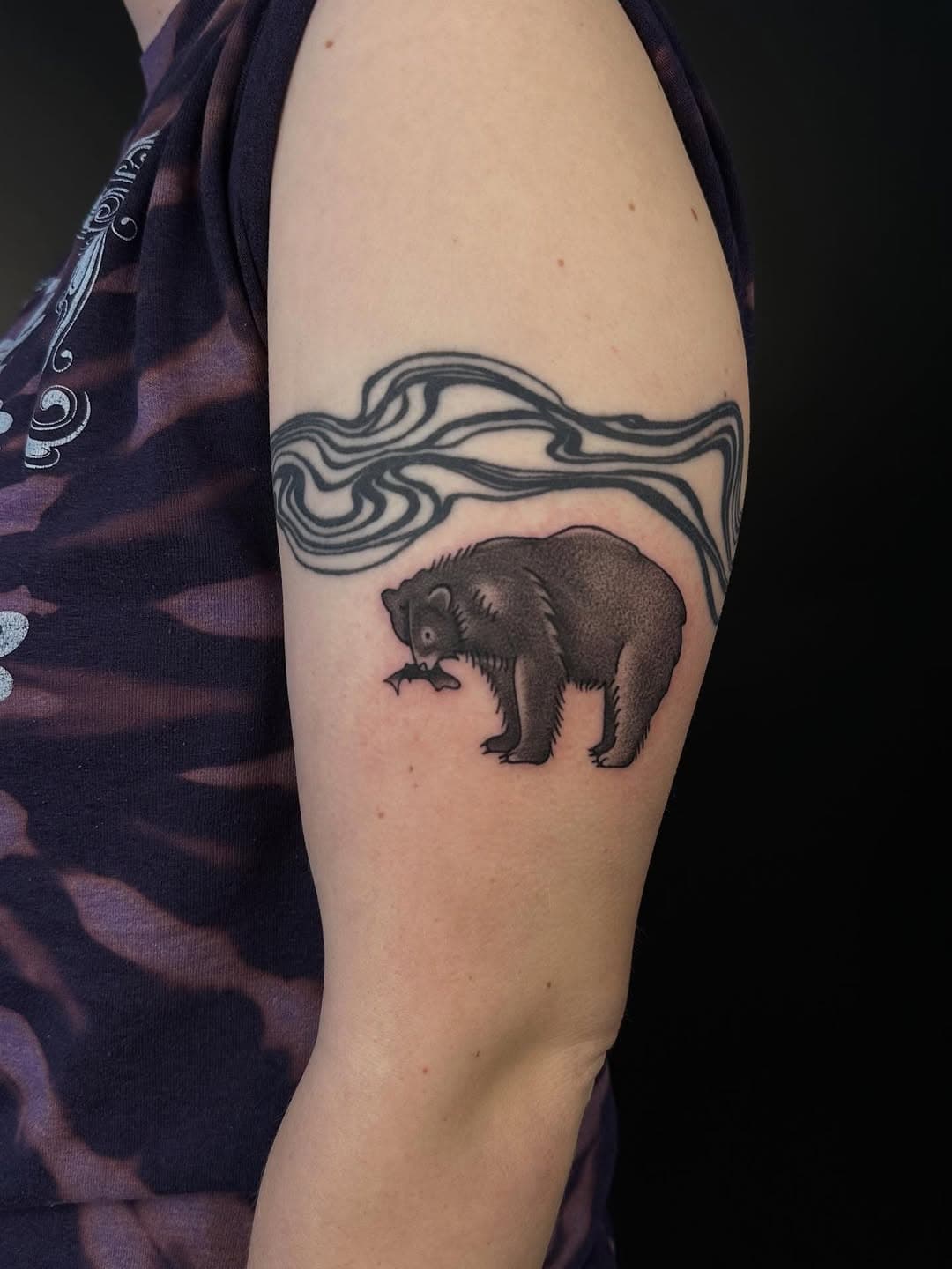Black and grey illustrative tattoo of a bear with flowing topographic line waves above it on upper arm