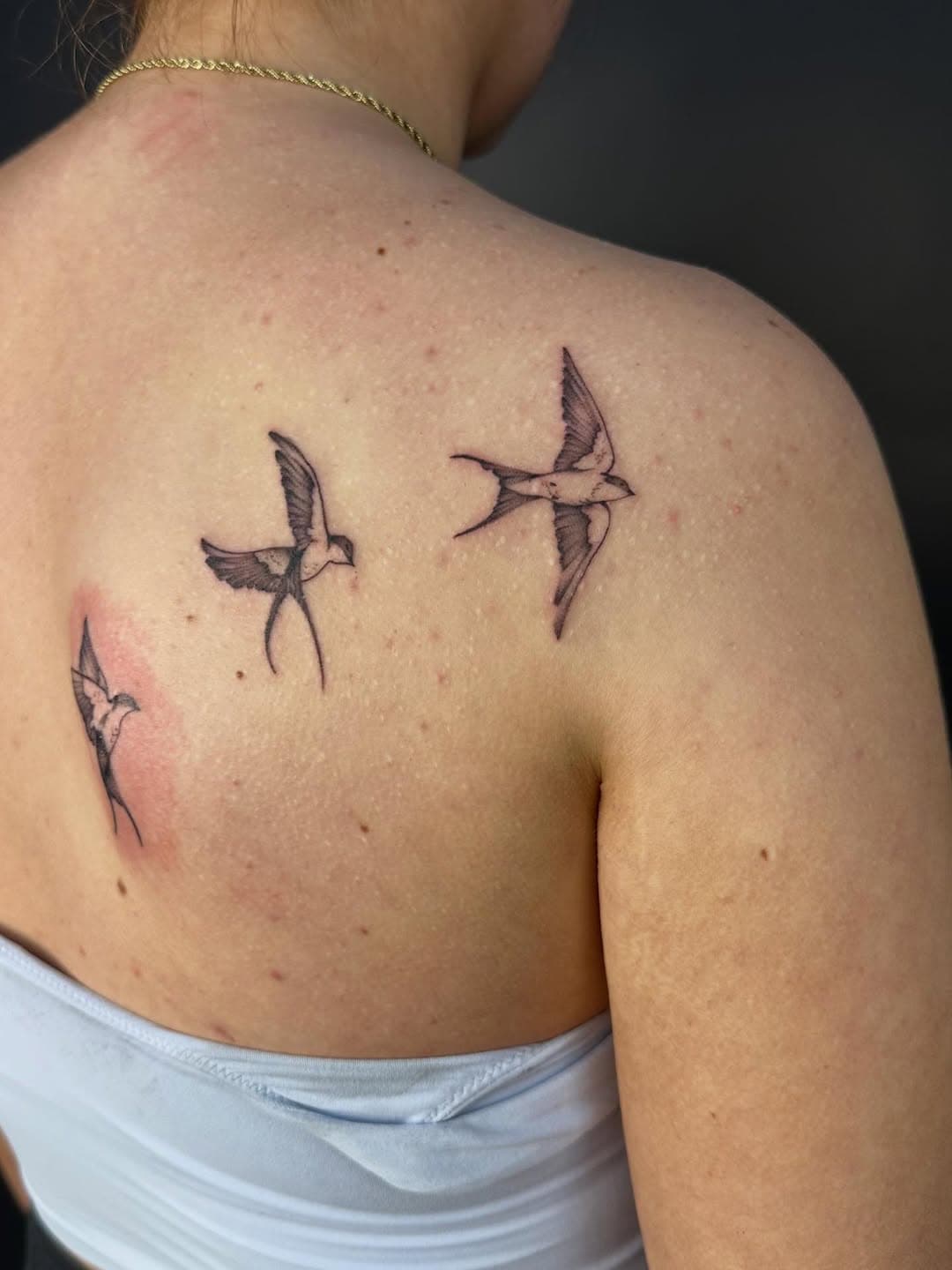 Three delicate black and grey swallows in flight tattooed on upper back and shoulder in fine-line illustrative style
