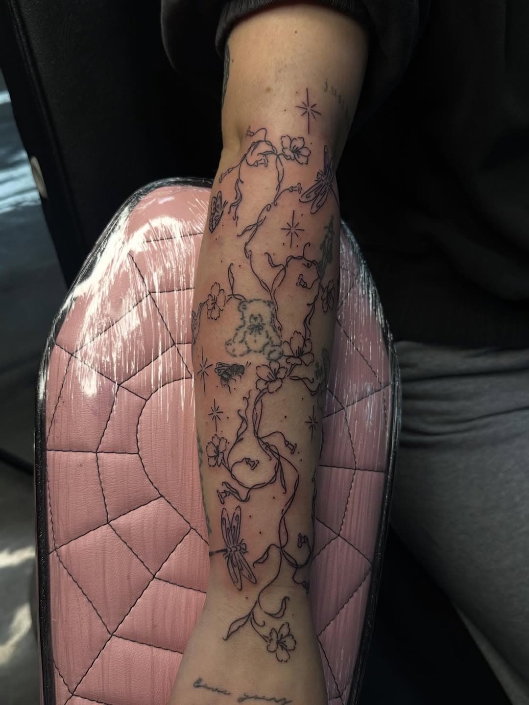 Delicate black line tattoo of flowering vines with stars wrapping around forearm, resting on pink quilted chair arm