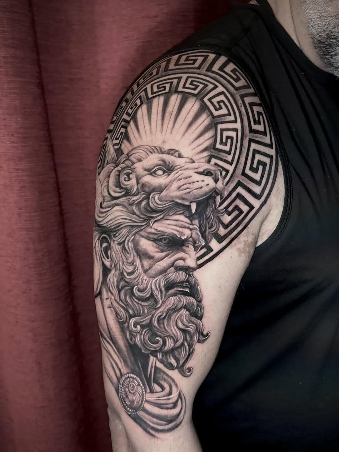 Detailed black and grey shoulder tattoo featuring Hercules wearing Nemean lion headdress with Greek key pattern border and radiating lines