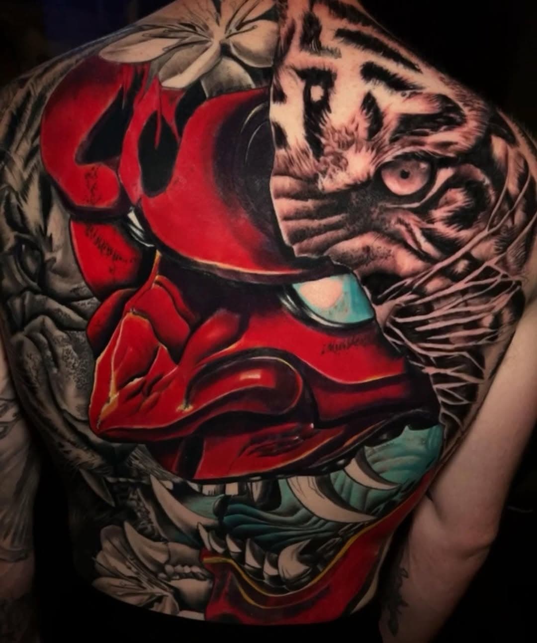 Full back tattoo featuring a red demon mask merged with a realistic tiger face, surrounded by flowers in neo-traditional style