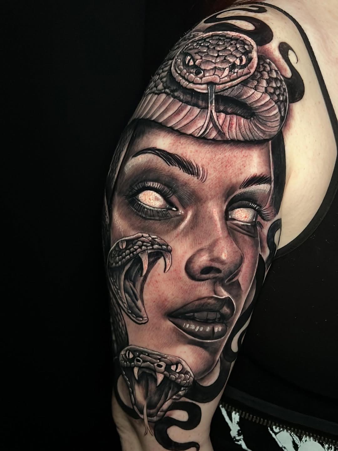 Neo-traditional tattoo of a woman's face with blank white eyes surrounded by coiled serpents with open fangs in black and grey realism