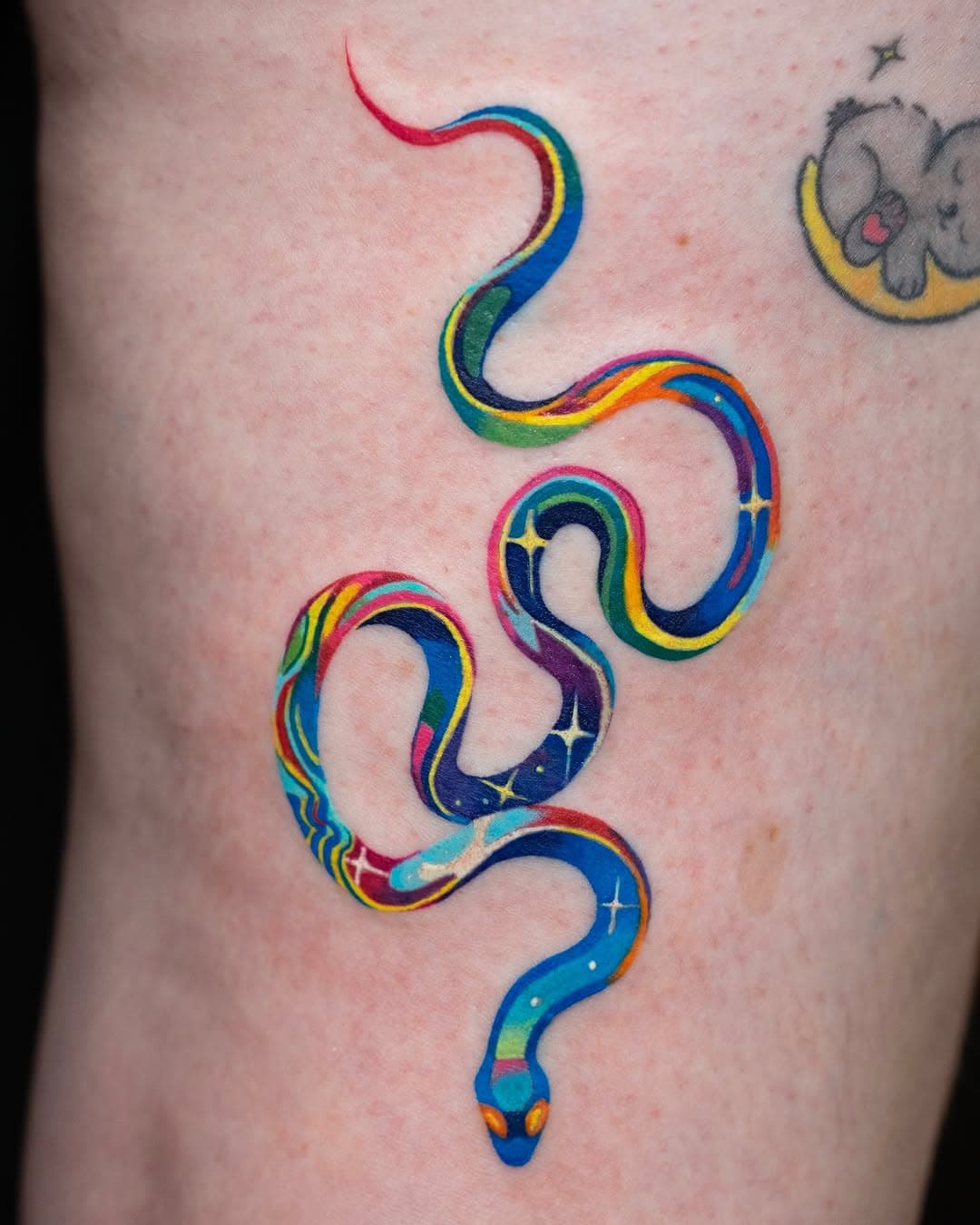 A vibrant rainbow-colored snake tattoo with cosmic star details winding down the body in an illustrative watercolor style