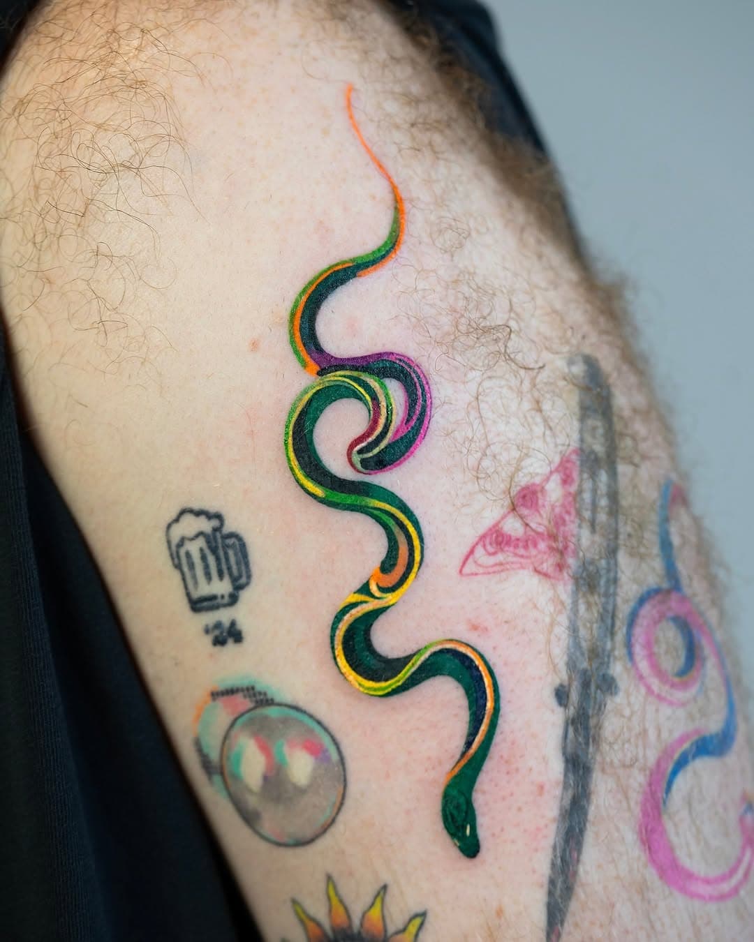 A colorful snake tattoo with rainbow gradient stripes winding down an arm, surrounded by other tattoos including a beer mug and circular designs