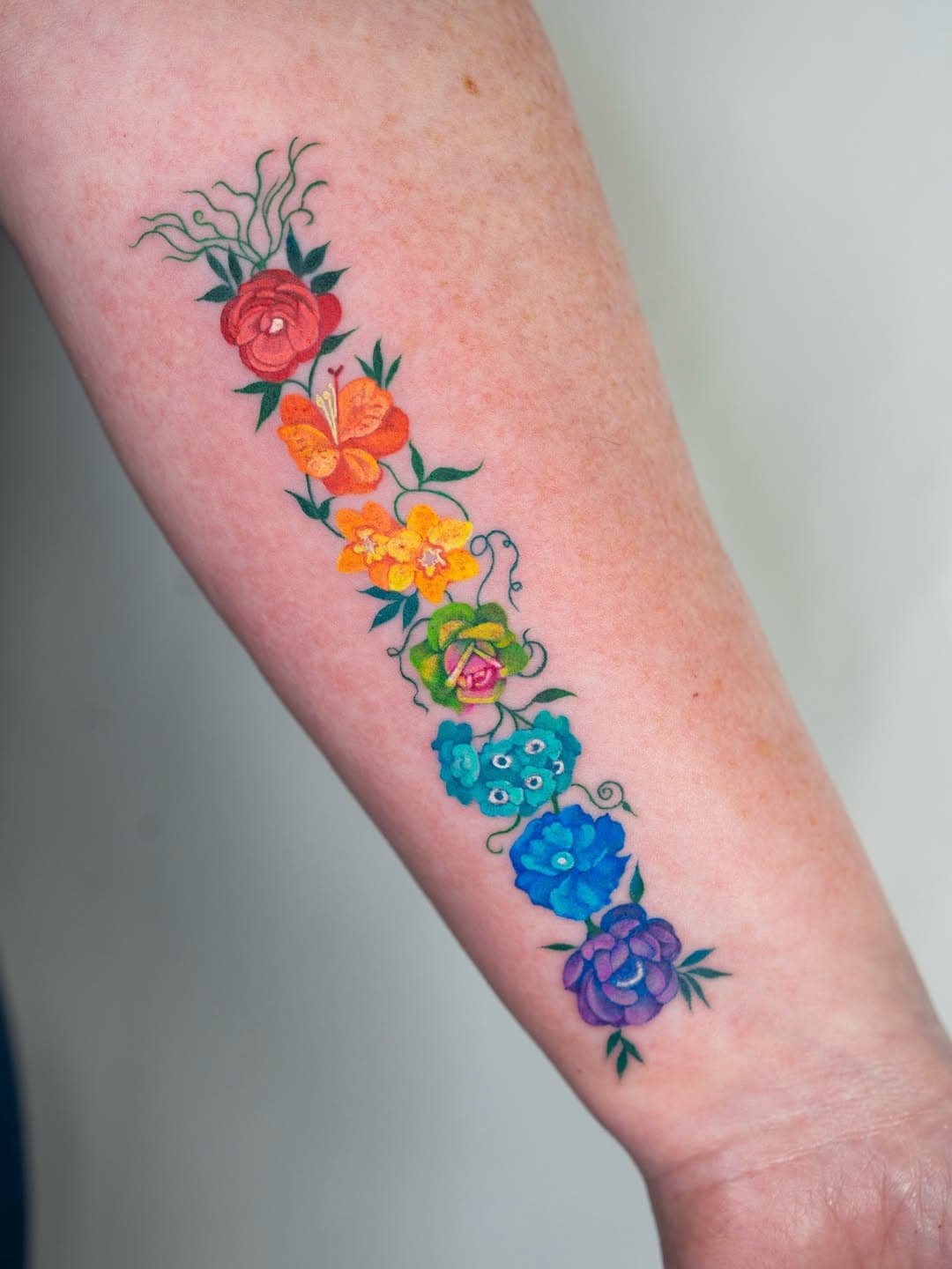 Rainbow-colored floral tattoo arranged vertically on forearm, featuring vibrant flowers in red, orange, yellow, green, blue, and purple hues