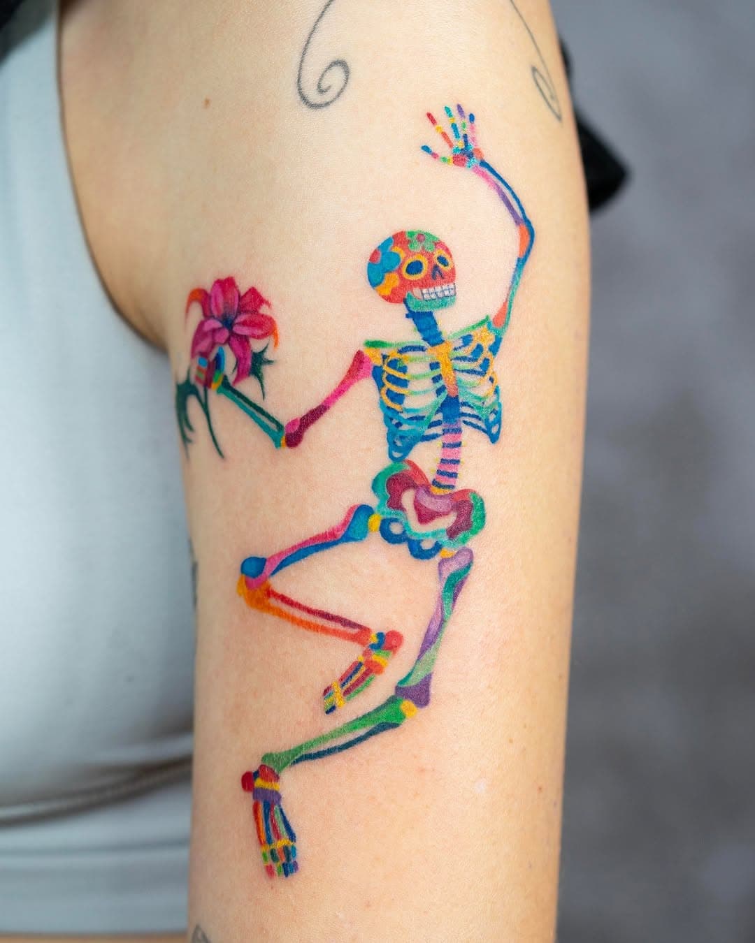 Colorful watercolor skeleton tattoo in dancing pose holding flowers, featuring vibrant rainbow hues throughout the bones and skull