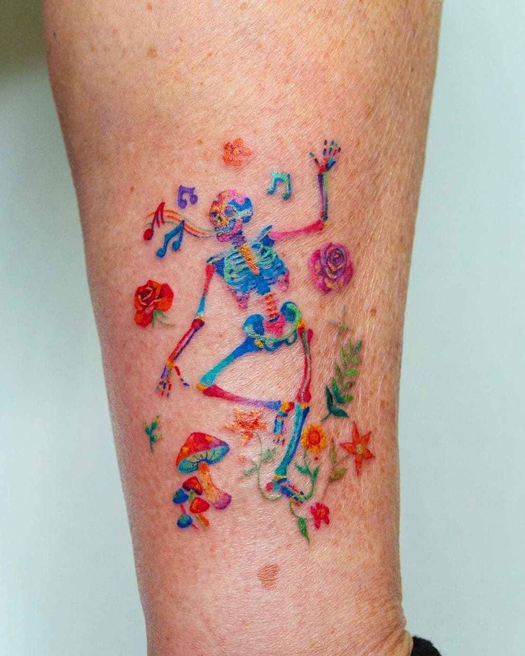 Colorful watercolor skeleton tattoo dancing among musical notes, roses, mushrooms and flowers on forearm