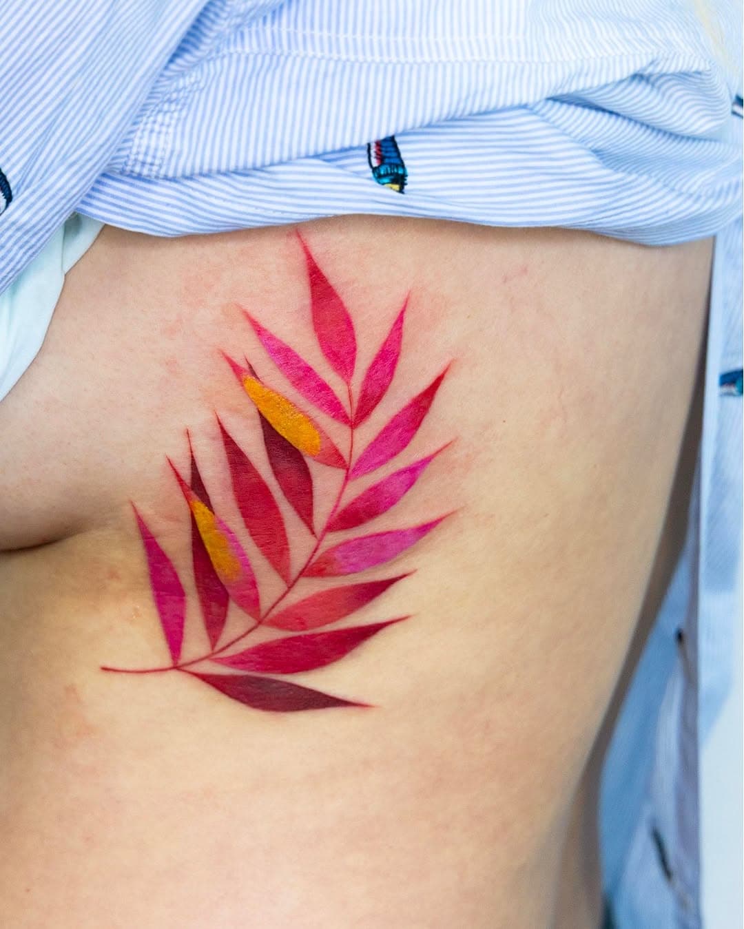 Vibrant watercolor tattoo of a tropical palm leaf in shades of red, pink, orange and burgundy on person's ribcage