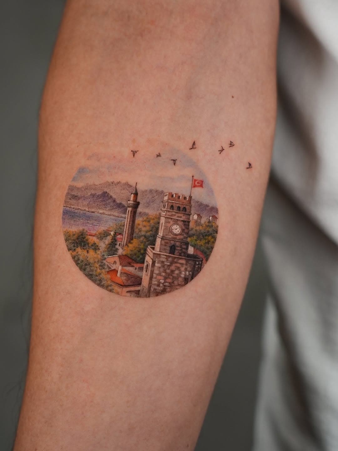 Circular illustrative tattoo of Turkish cityscape featuring clock tower with flag, minaret, waterfront, mountains, and flying birds on forearm